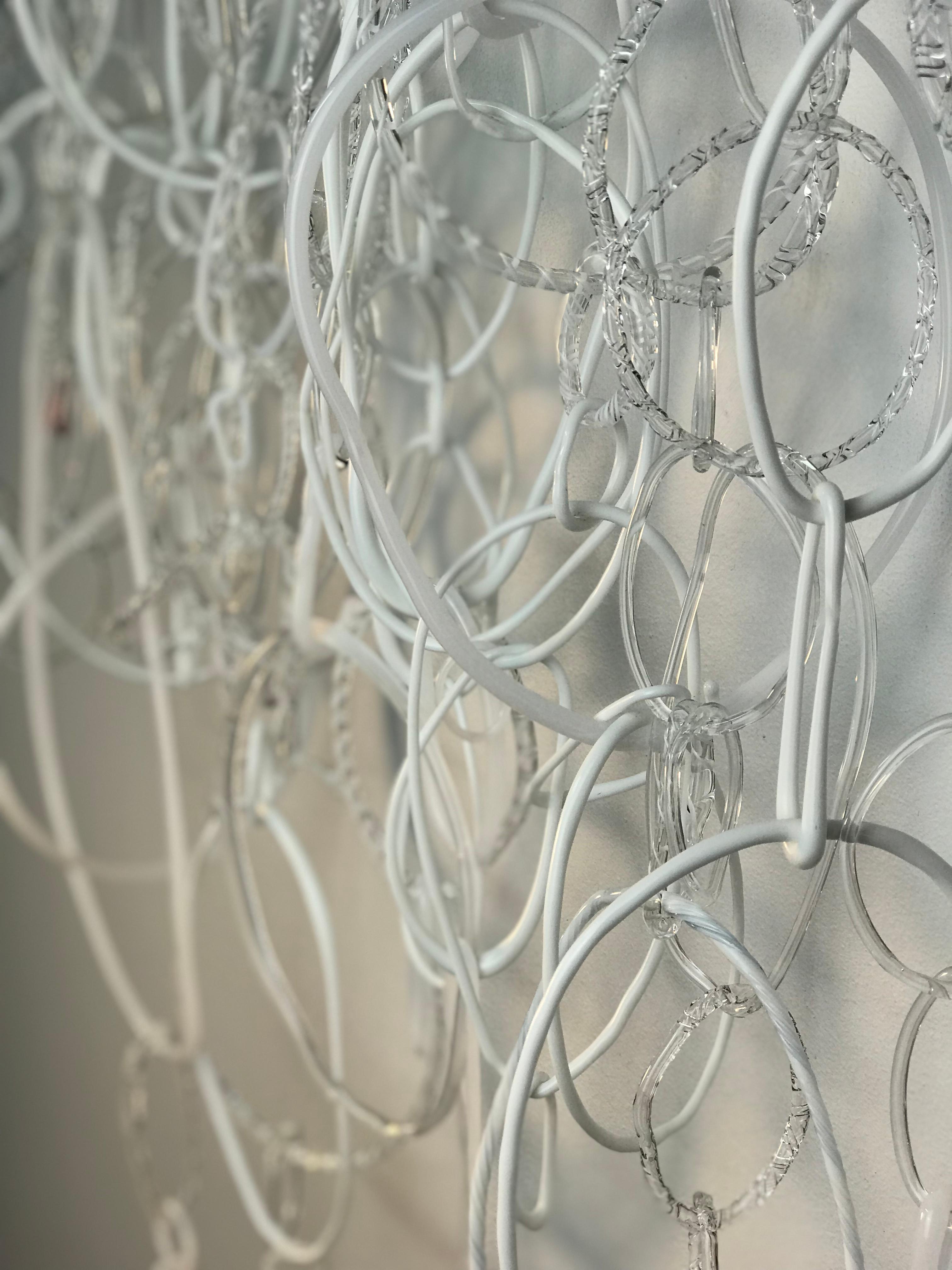This hanging sculpture is composed of freeform circular loops in various shapes and textures of white and clear torch worked borosilicate glass, linked together and hanging from an irregularly shaped translucent white glass rod supported by