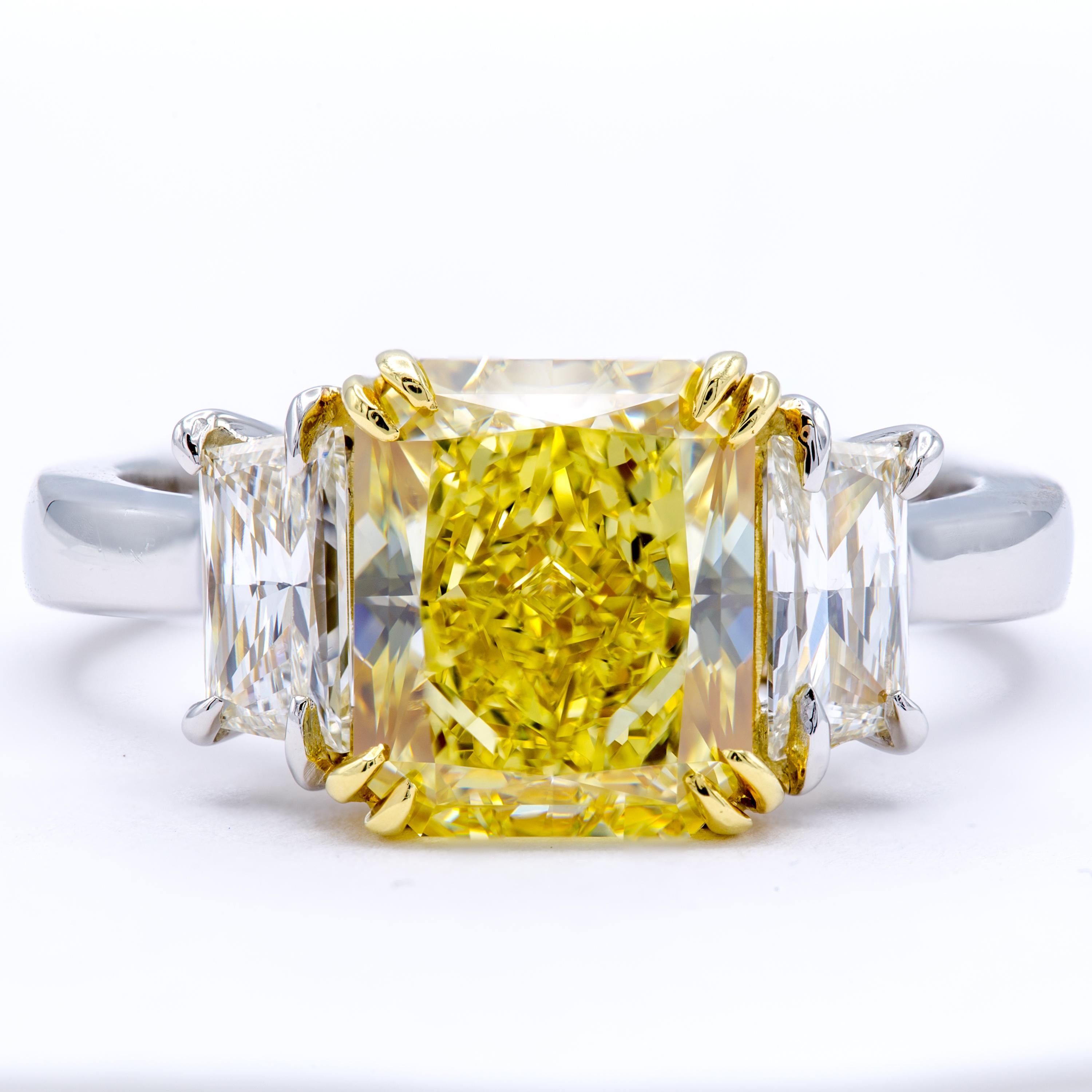 A platinum band wraps gently around the finger, securing an exceptional GIA certified 3.15 carat natural fancy intense yellow radiant diamond within a cradle of 18kt yellow gold. Two brilliant-cut trapezoid side stone diamonds accent wonderfully on