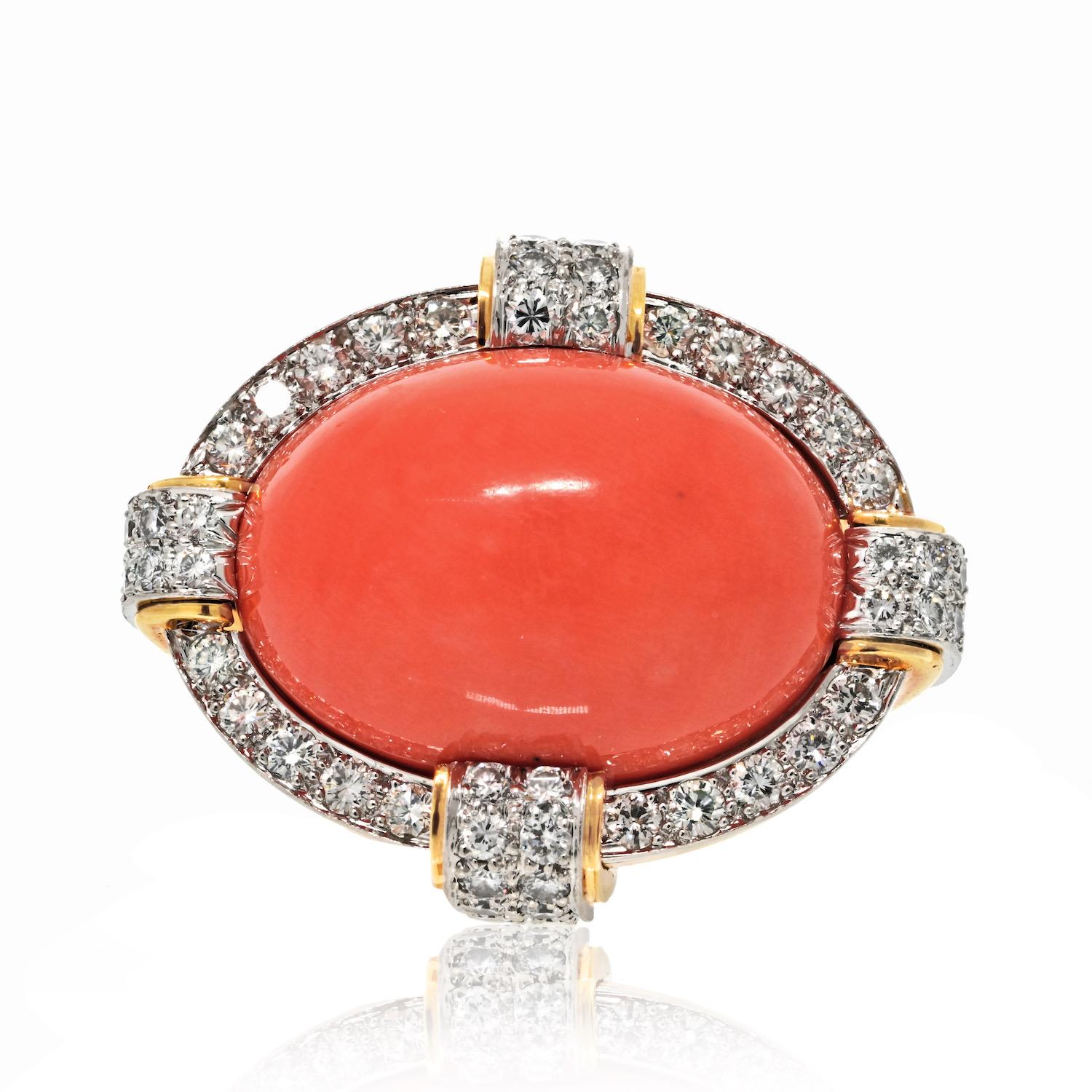This David Webb 18K Yellow Gold and Diamond Cocktail Ring is a bold and luxurious piece, perfect for making a statement. At its center, a striking oval cabochon-cut coral takes the spotlight, surrounded by a halo of brilliant round-cut diamonds. The