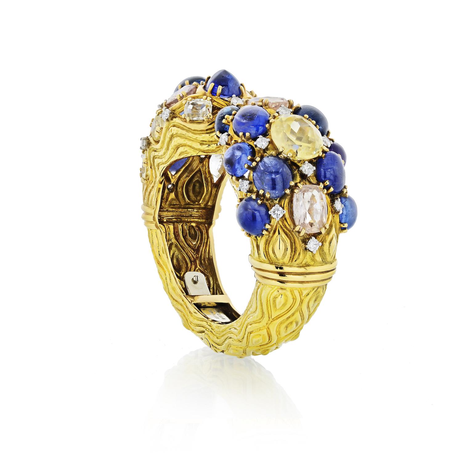 This bold yellow gold cuff bangle by David Webb features natural blue sapphires. Sculpted in 18k yellow gold as a hinged cuff you will be making a bold statement wearing this accessory. 
Set at the top with oval cabochon sapphires and circular-cut