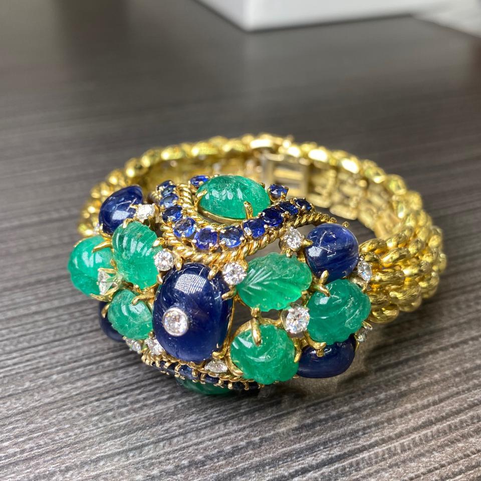 David Webb Carved Emerald Sapphire Diamond Bracelet.
This stunning David Webb bracelet is set with circular-cut and cabochon sapphires, brilliant-cut diamonds and emeralds carved with foliate motifs, to a strap of textured links, inner circumference
