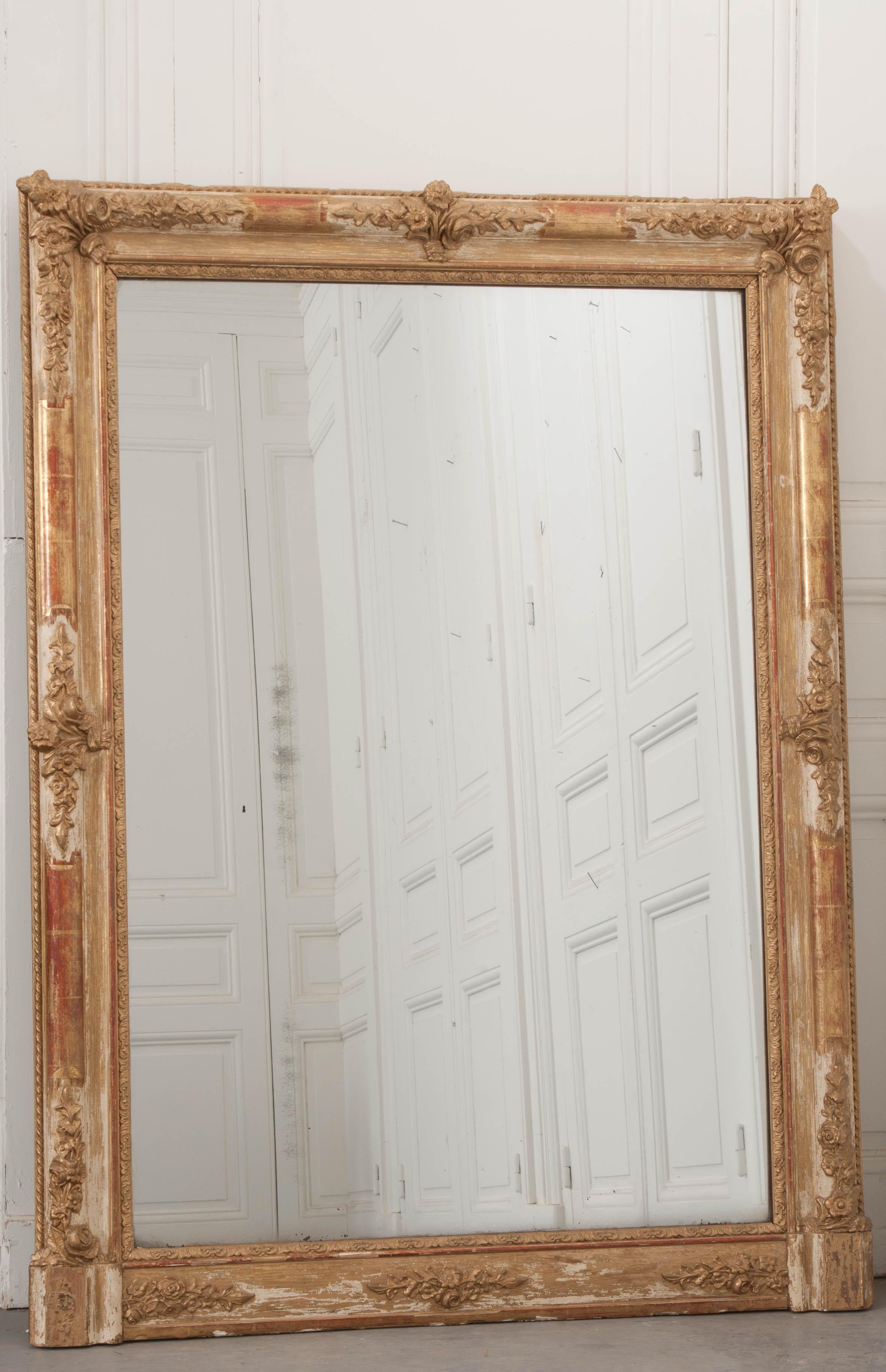 A brilliant 19th century giltwood over-mantle mirror, made in France circa 1870. The mirror is large and beautiful. The frame is ornamented with bundles of flowers that can be found in its corners and sides. The gilt finish has experienced some loss