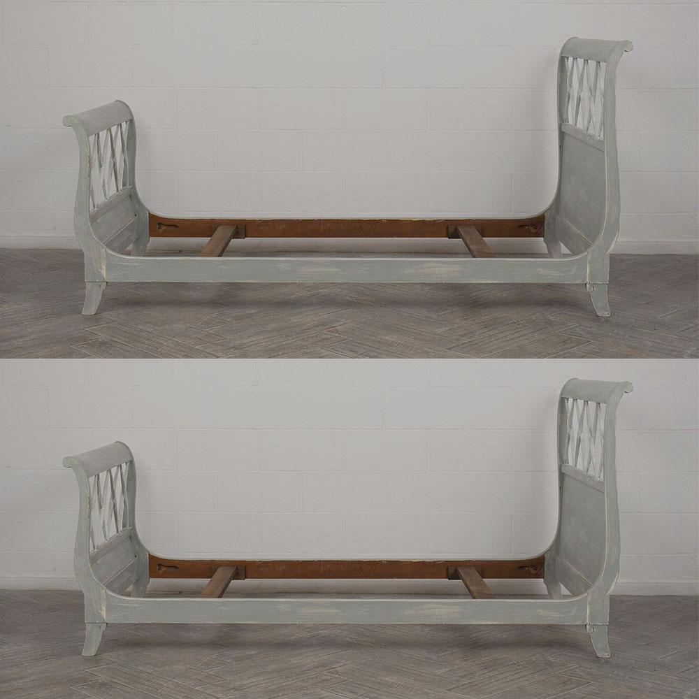 This pair of 1950s Empire-style bed frames feature a carved wood frame that has been painted in a pale grey and off white color combination with a distressed finish. The carved frames are very elegant design with scroll arms and X-design on the