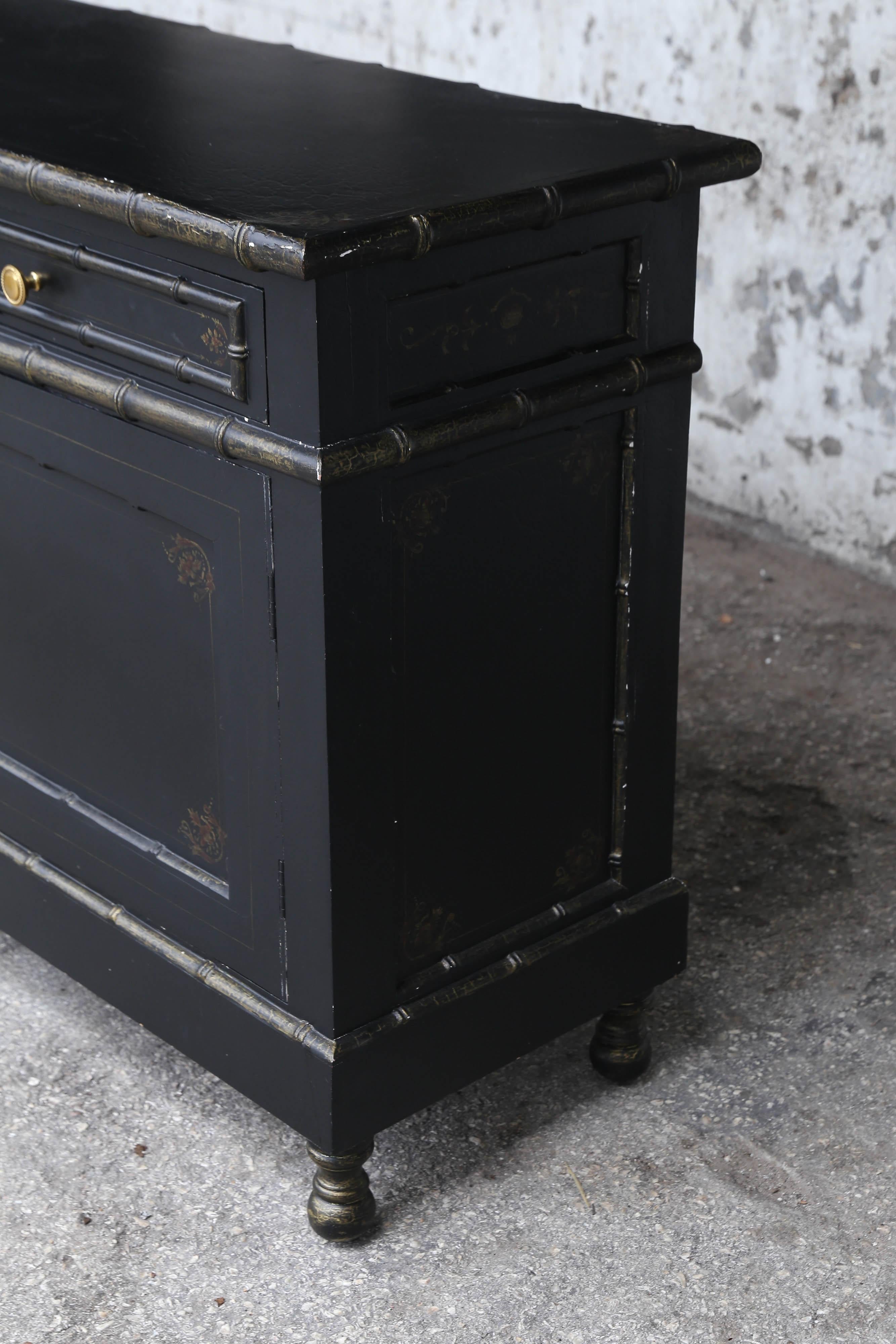 Vintage faux bamboo black painted credenza features two doors and two drawers with gold paint accentuating the shapes of bamboo.
Faint gold patterns are painted on credenza top and front of two doors.
Credenza sits upon 4 low turned legs and has