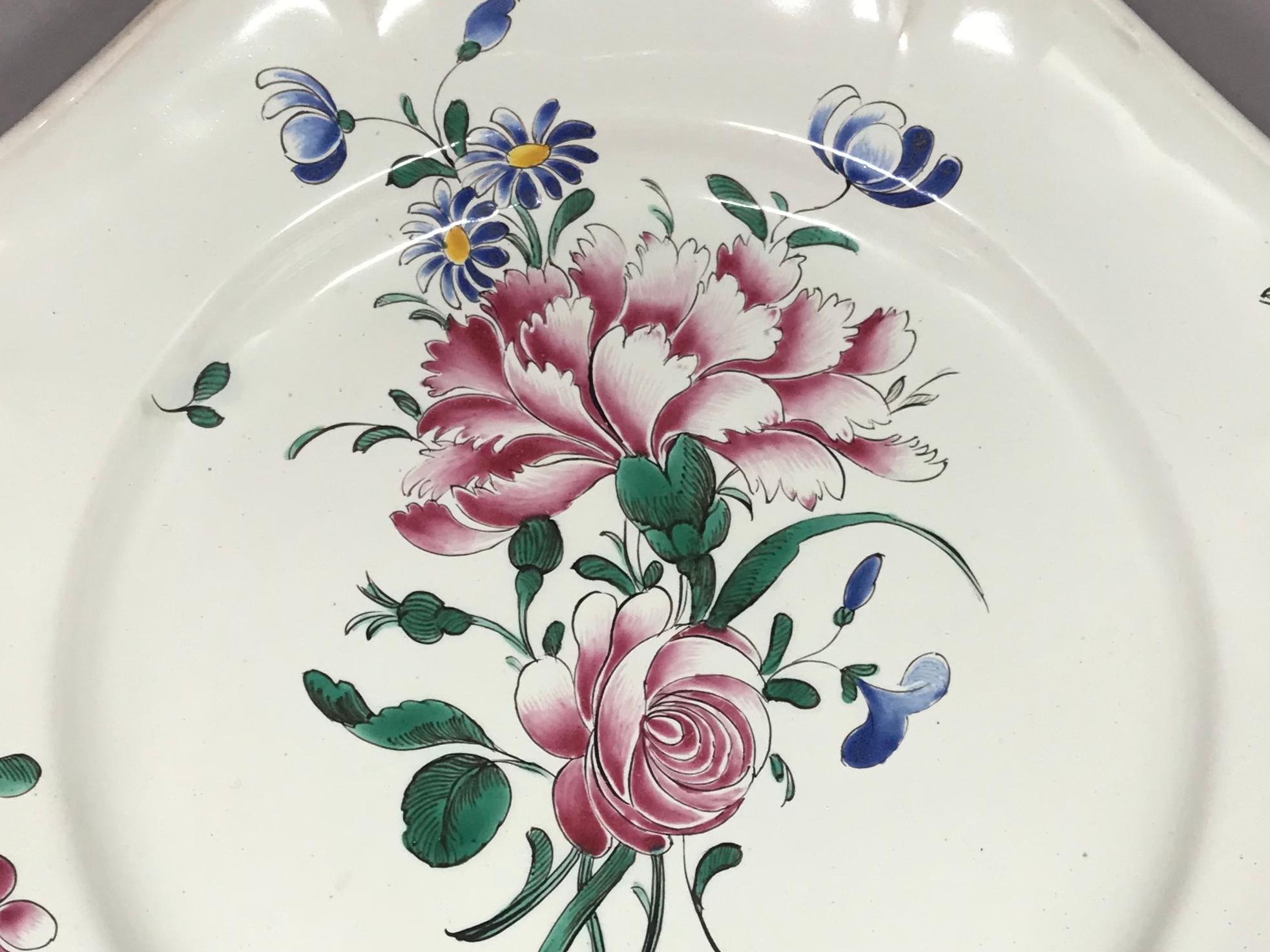 Plaque florale en faïence de Strasbourg. Assiette ancienne en faïence avec un grand bouquet floral de roses et de bleus, avec un bord lobé et d'autres gerbes florales ; avec des marques sous glaçure bleue pour la manufacture Hannong, France, vers