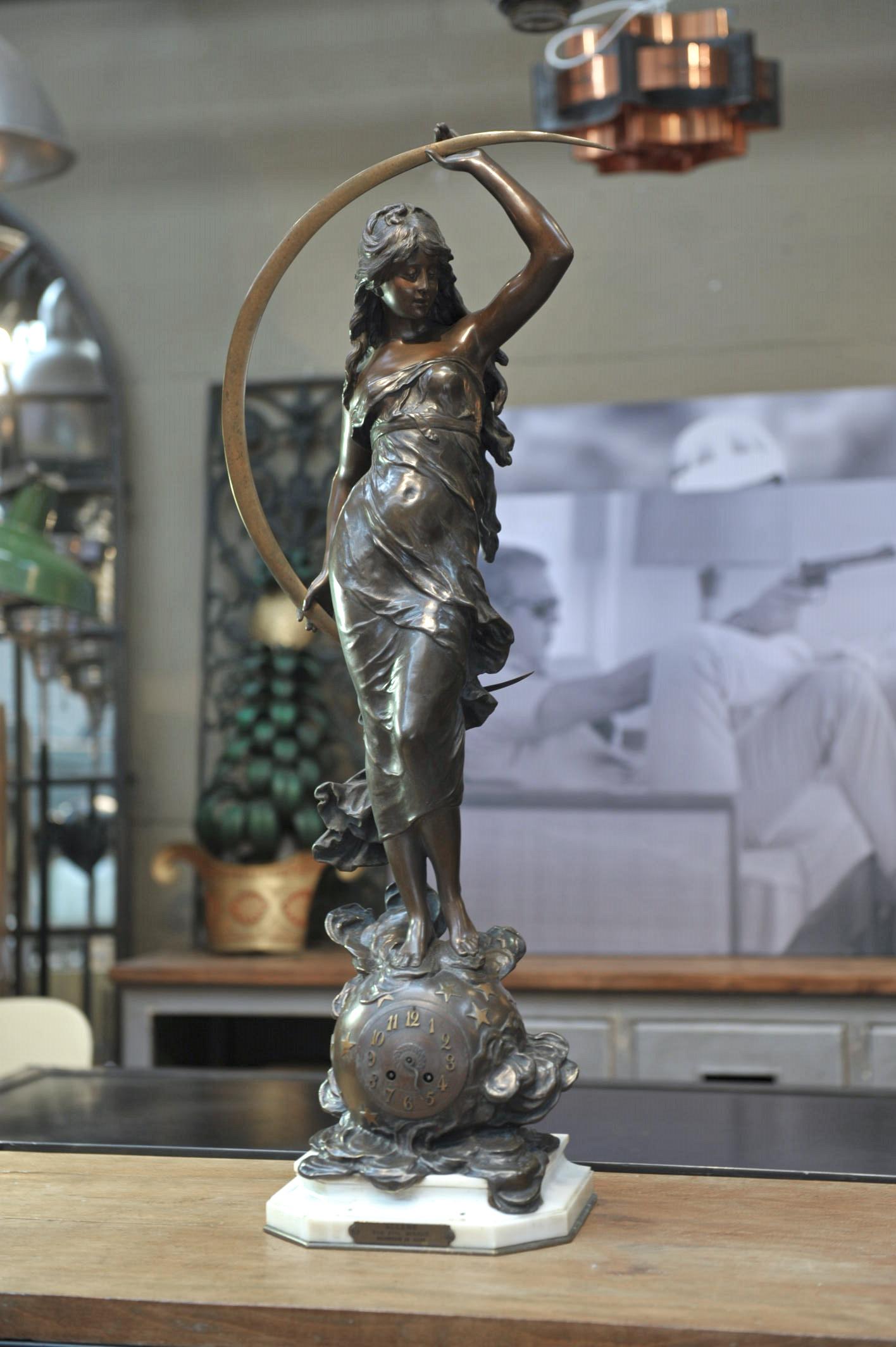 Bronze sculpture with brown patina including a clock and signed Auguste Moreau. It features a woman wearing a flowing dress who is standing on a globe and holding a waning crescent moon in her hands. The title printed on a small plaque reads:
