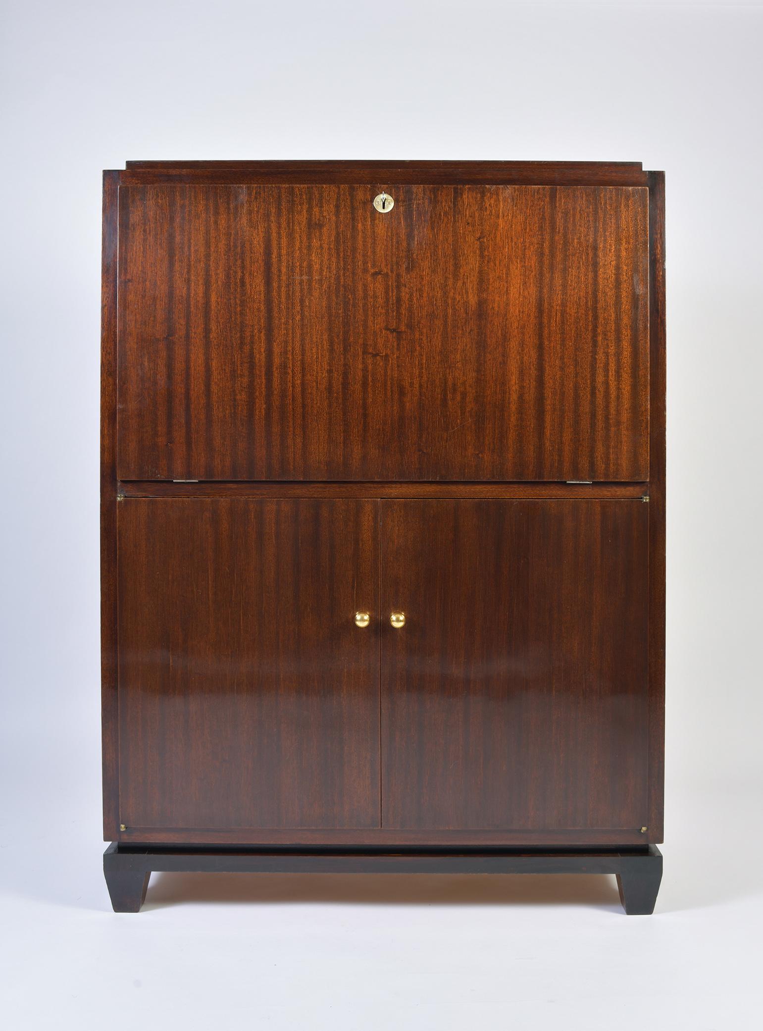 An Art Deco mahogany cabinet, the top section opening with an abattant which can used either as a secretaire or as a bar, locked with a brass key hole and its key in working order, the lower section opening with two doors with solid brass knobs,