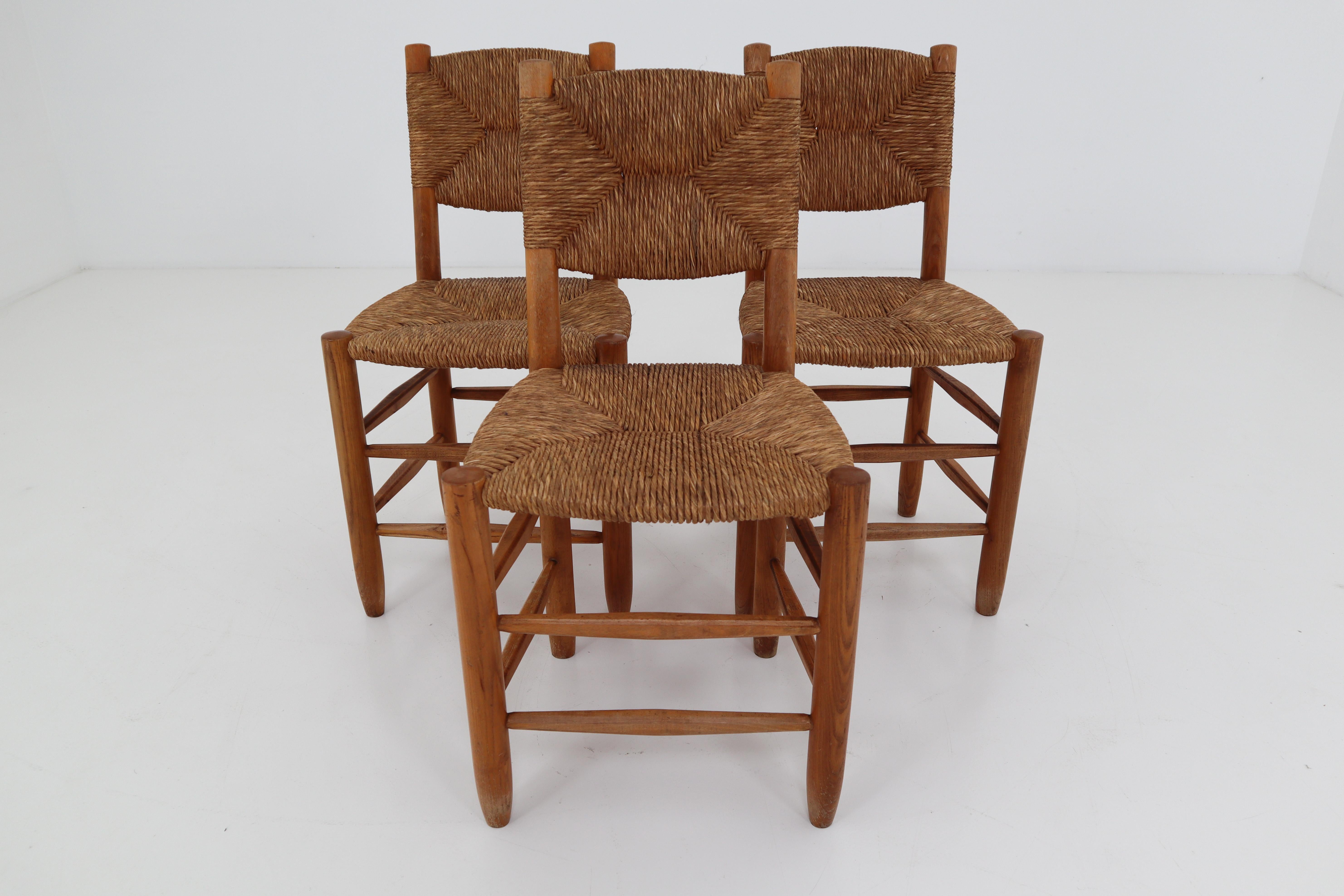 A pair of three dining chairs of wood and straw in one of the quintessential midcentury designs of their creator, the famous French furniture maker Charlotte Perriand. The three chairs are identical in the patina as in their very uniform state.