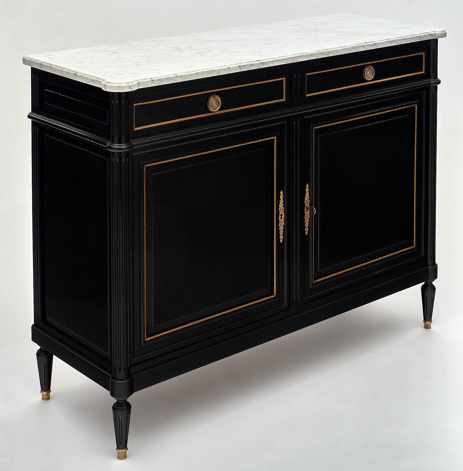 A flawless two-door French antique marble-topped buffet in the Louis XVI style. It is made of ebonized mahogany with a lustrous French polish finish. This piece has two dovetailed drawers above two doors, all featuring gilt brass trims and hardware.