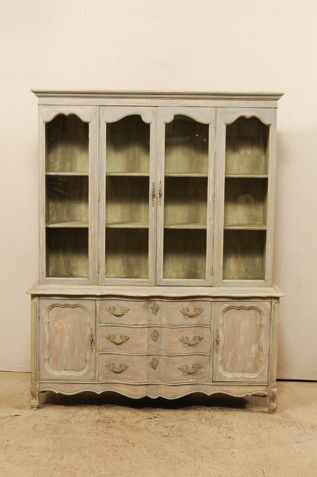 A tall American glass and painted wood cabinet from the mid-20th century. This vintage cabinet, created in the French style, has a glass display top with two doors, atop a bottom cabinet with three central drawers flanked with two wood doors at