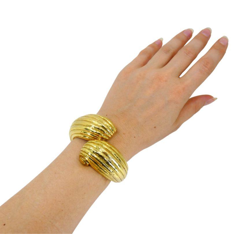 Ce bracelet manchette David Webb des années 1990 est une pièce robuste, principalement fabriquée en or 18 carats avec des éléments en platine pour sa fermeture mécanique à charnière. Le design du bracelet présente un motif croisé en forme de