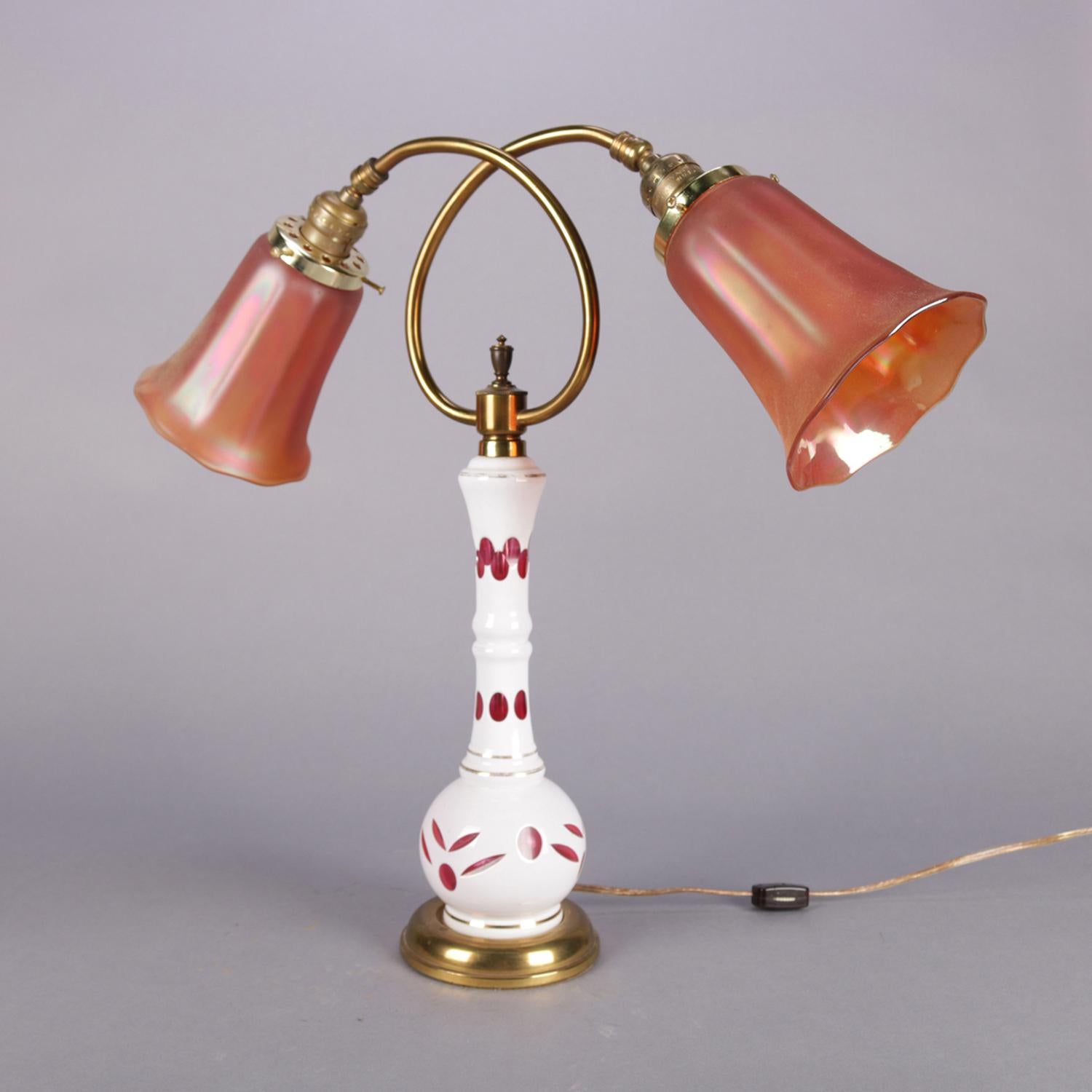 Antique Bohemian glass table lamp features cased cut to cranberry vase with stylized floral motif seated on brass base and having two scrolled arms terminating in lights with carnival glass faceted shades, professionally re-wired, circa