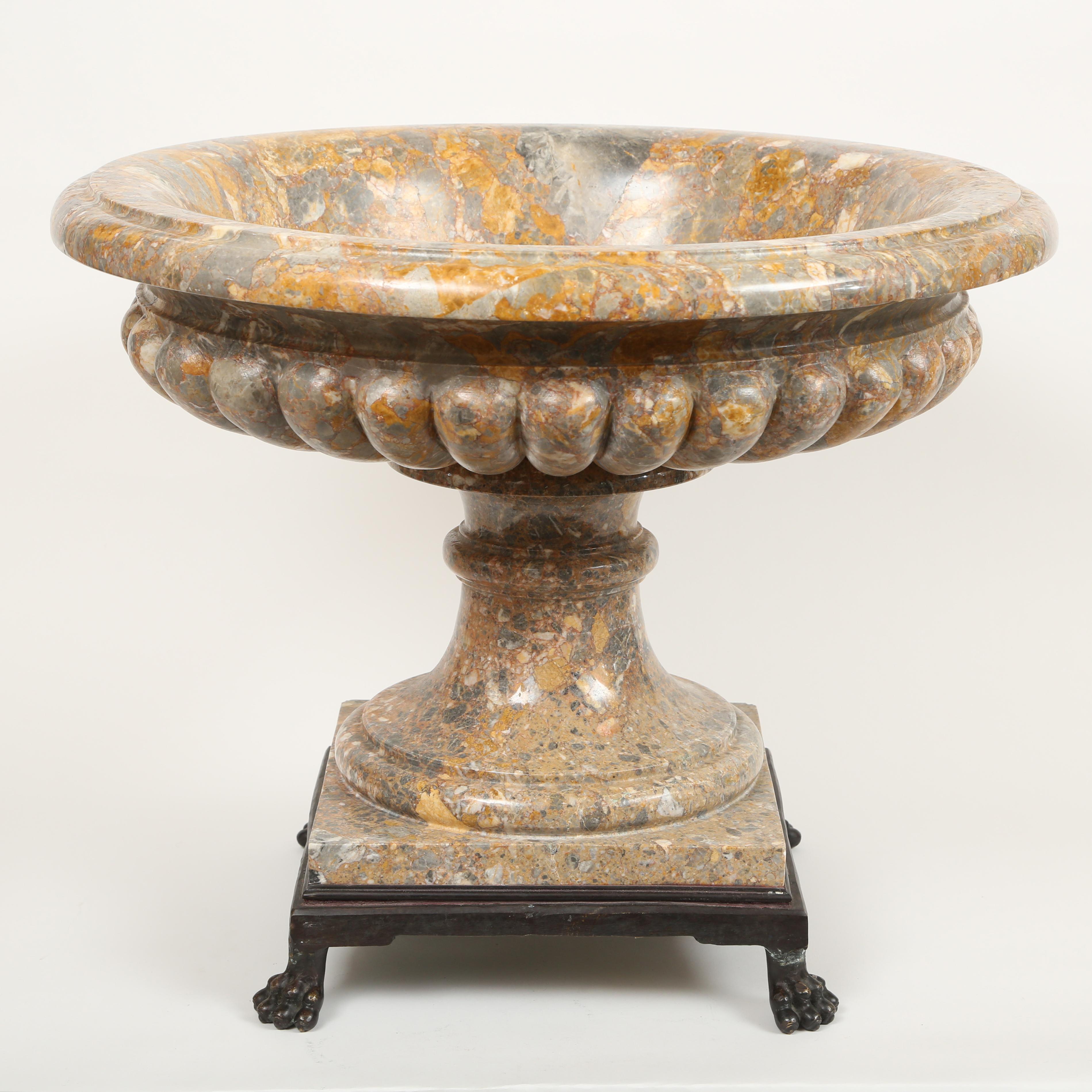 Large neoclassical wide mouthed tazza style shallow marble urn. The urn with everted rim has lobed or ribbed sides. The bowl of the two part urn sits on a circular socle and square base. The entire piece fits into an iron base with lion's paws.
The