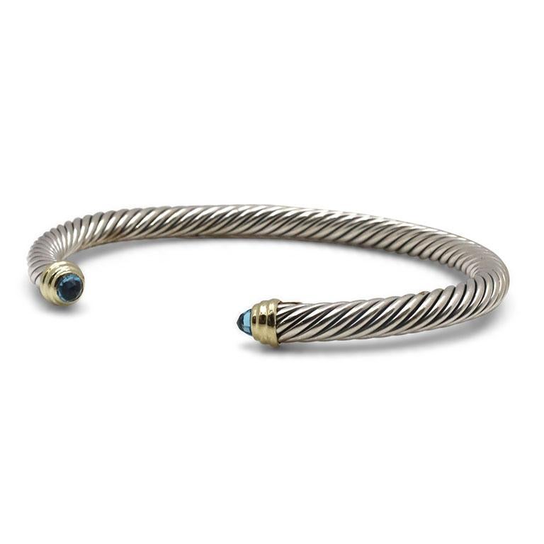 Authentic David Yurman Cable bracelet crafted in sterling silver with 14 karat yellow gold accents and featuring two blue topaz stones. This classic bracelet measures 5mm in width and can fit up to a size 6 1/2 inch wrist. Signed DY, 585 and 925.