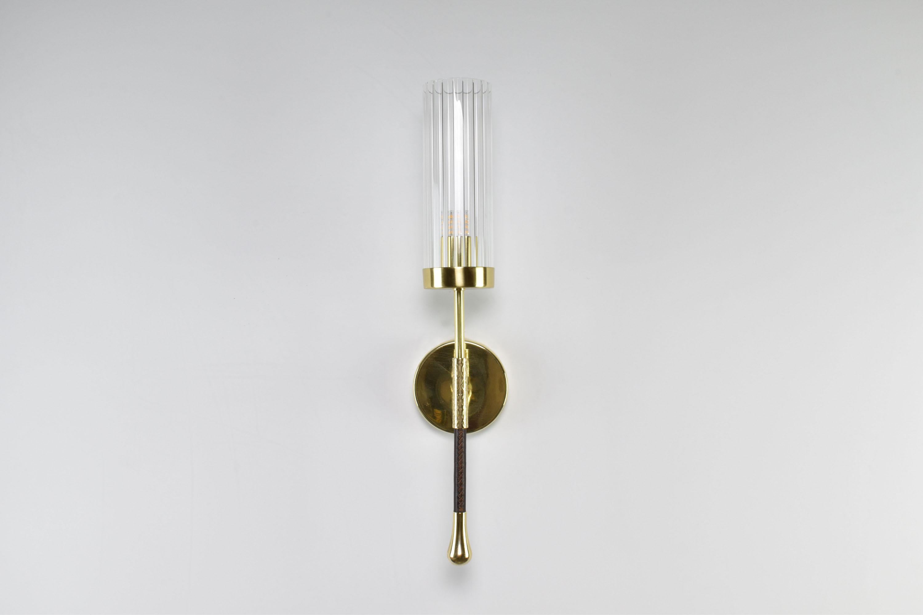 The Daya-wo1 wall light boasts a timeless brass structure. Its leather-sheathed stem at the bottom adds a touch of refinement to the overall design, while the cylindrical textured glass shade provides a subtle yet stylish illumination to any space.