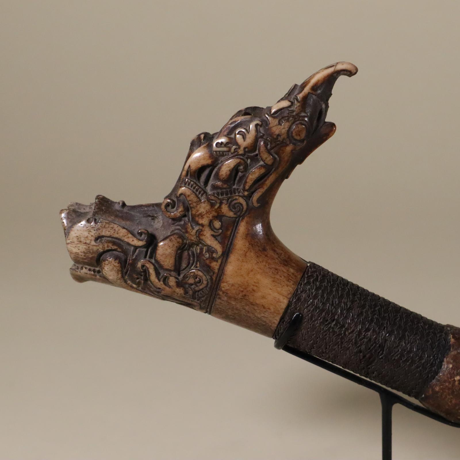 This is a handle for a sword (mandau) from the Dayak (var. Dyak) people of northern Borneo. Carved from deer antler. A grip of plaited fiber covers the shaft. The handle has a long spur on one side and a short spur on the other. The whole is adorned