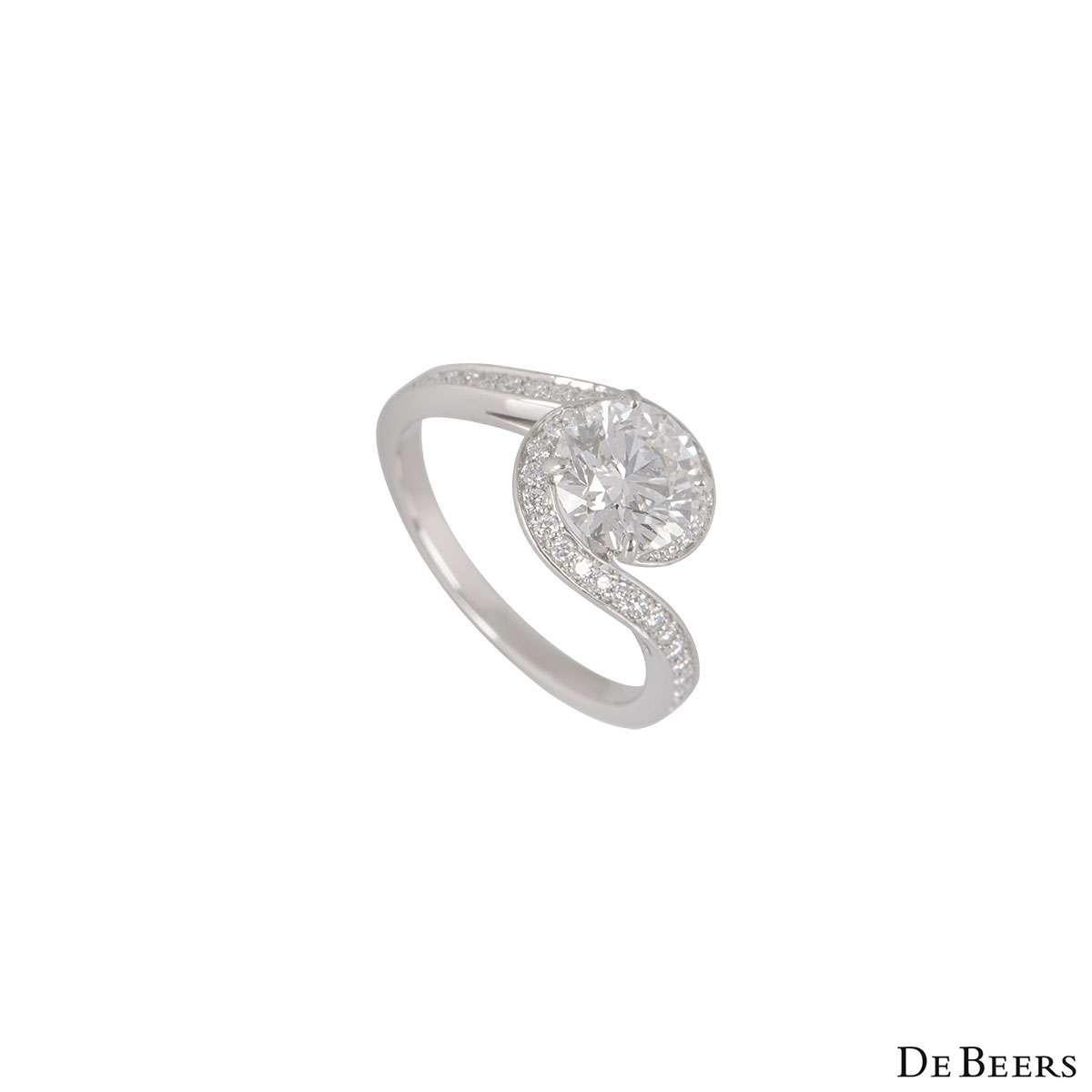 A stunning platinum diamond ring by De Beers from the Caress Collection. The ring features a round brilliant cut diamond within a four claw setting with a weight of 1.24ct, H in colour and SI1 clarity with a swirl of pave set diamonds around it