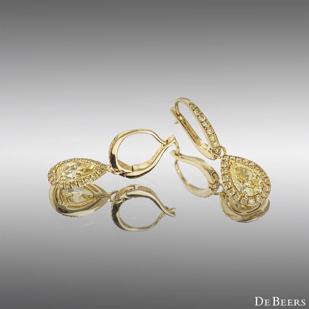 An elegant pair of 18k yellow gold diamond earrings by De Beers from the Aura collection. The drop earrings each comprise of a half hoop with 8 round brilliant cut yellow diamonds suspending a pear shaped fancy yellow diamond and further