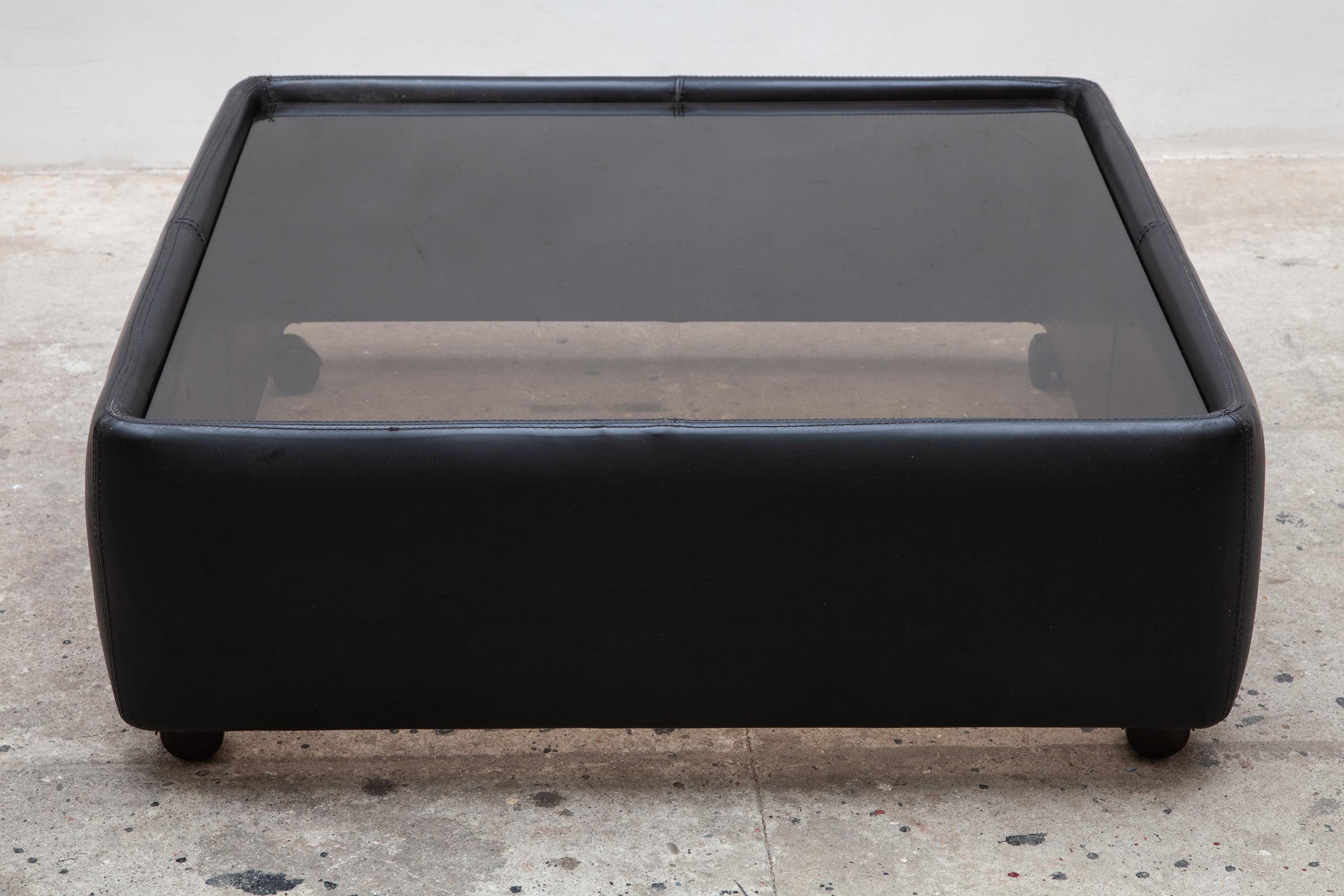 Minimalist 1970s entire piece is upholstered in black leather with a smoked glass top coffee or cocktail table by De Sede, Switzerland. Rests on four brown plastic casters.