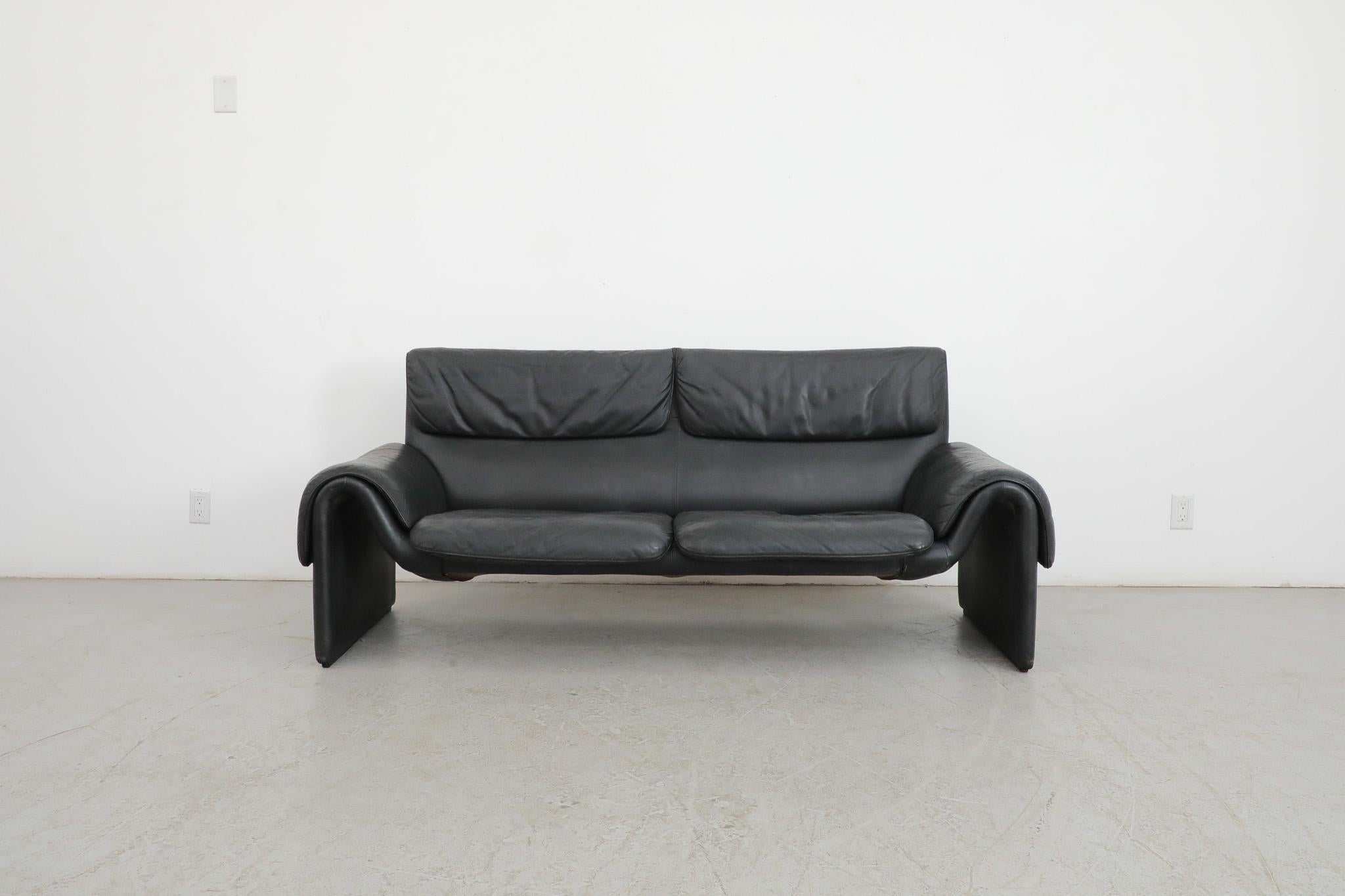 Handsome 1980's black leather De Sede 'DS2011' two seater sofa with dramatic swooping armrests that are integrated with its curvy frame. Sleek and super stylish sofa with lovely patina and some visible wear consistent with its age and use.  