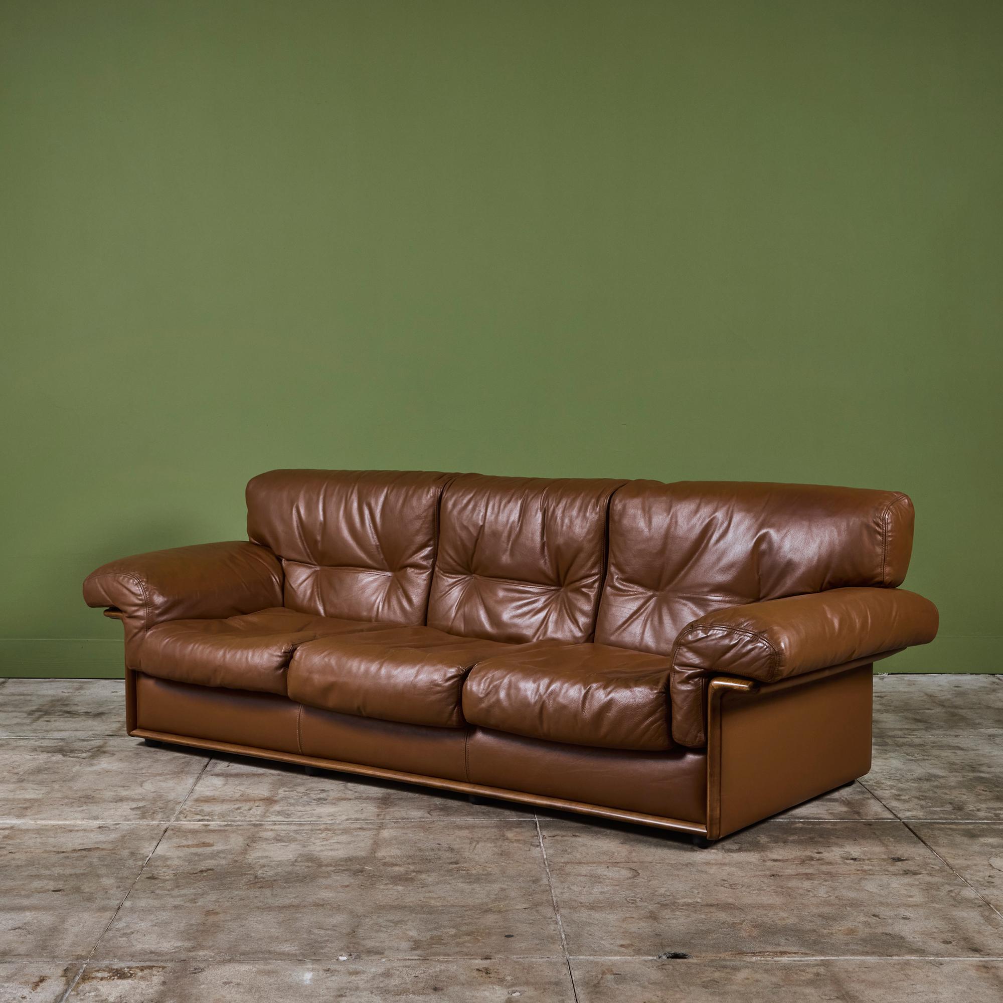 Three seat brown leather sofa in the style of De Sede. The sofa features a walnut frame with tufted seat backs and over sized cushions. The cushions and armrests showcase a double stitched detail. The entire sofa rests upon eight small peg