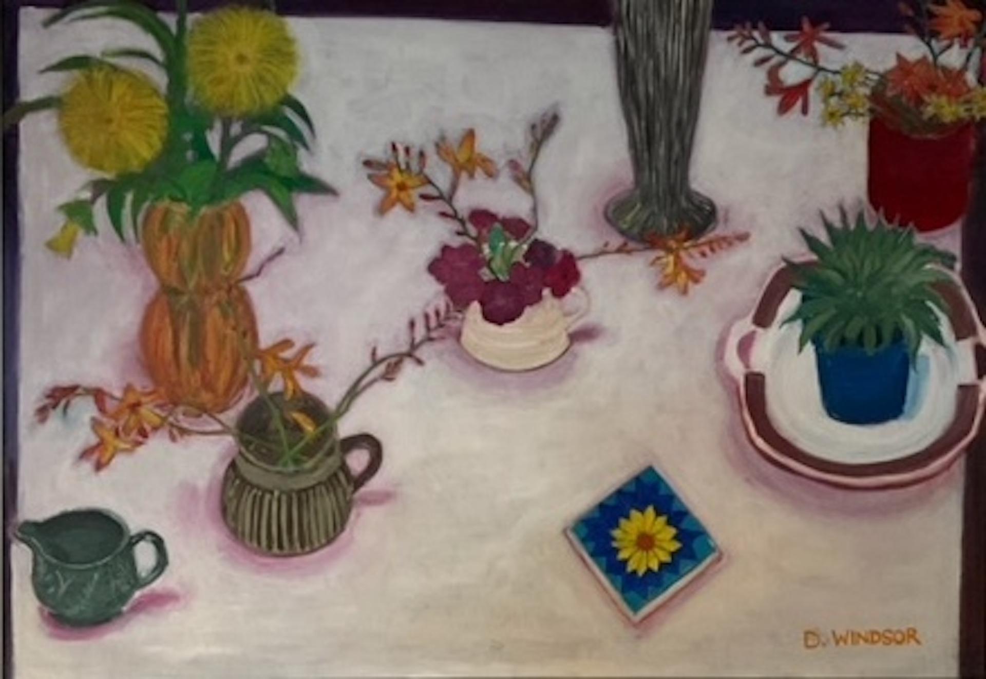 Nature morte sur nappe blanche" est une peinture originale de Deborah Windsor. Dans cette grande nature morte, de petits vases de fleurs d'été, un cactus et d'autres objets sont posés sur une nappe blanche impeccable. Placée dans un élégant cadre