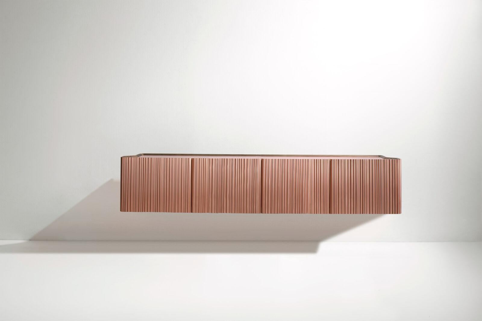 The sculptural surface is defined by fluting composed of vertical rods of different sizes in brushed metal, creating irregular undulations and a fascinating interplay of reflections. Extending horizontally, it has two L-shaped doors that open at 90