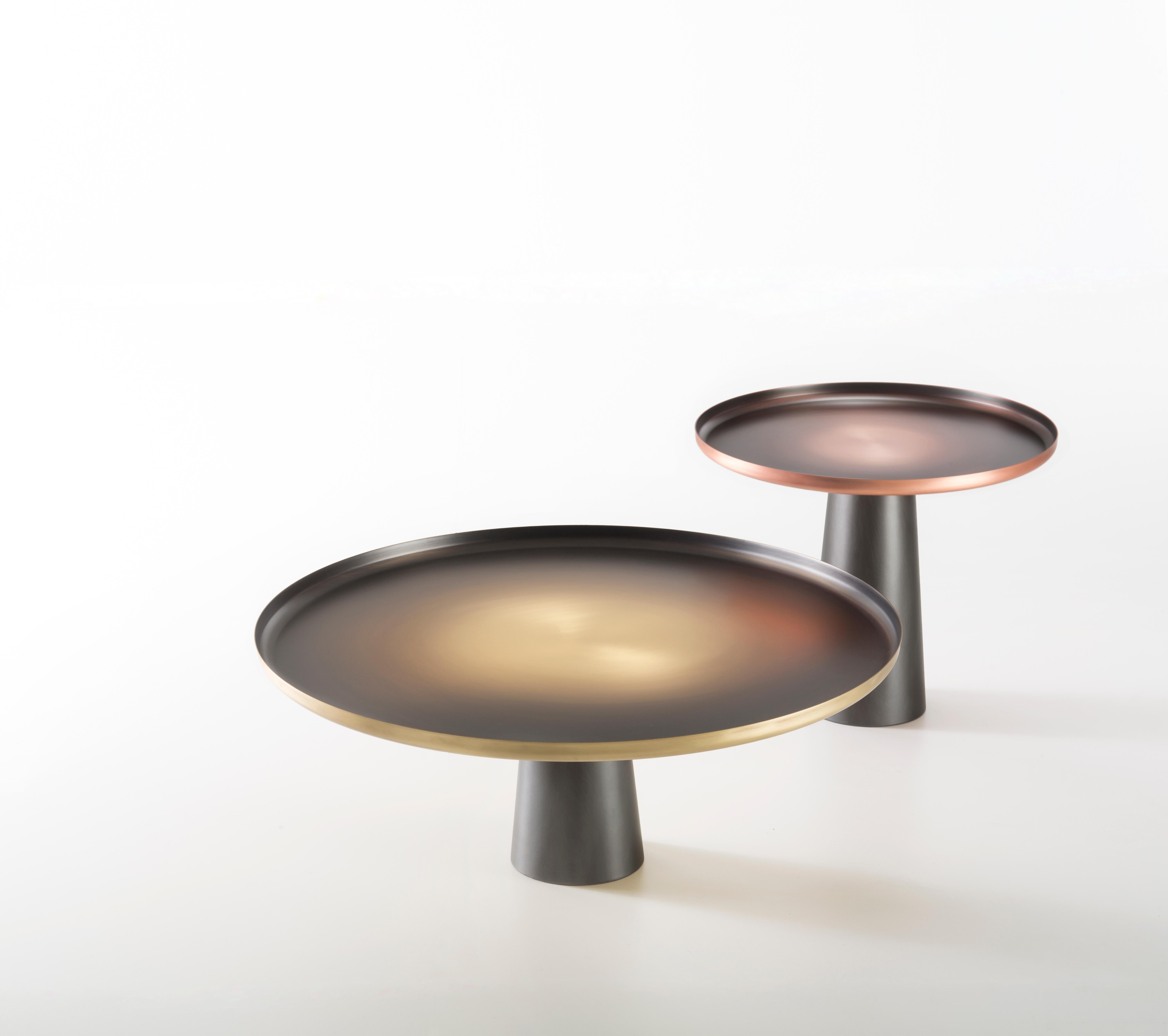 Two side tables, like large suns, emphasize the textural and chromatic characteristics of copper and brass. Contrasting with the DeLabré iron of the base, the top is ennobled by a hand-crafted finish. Here the material takes shape in all its purity,