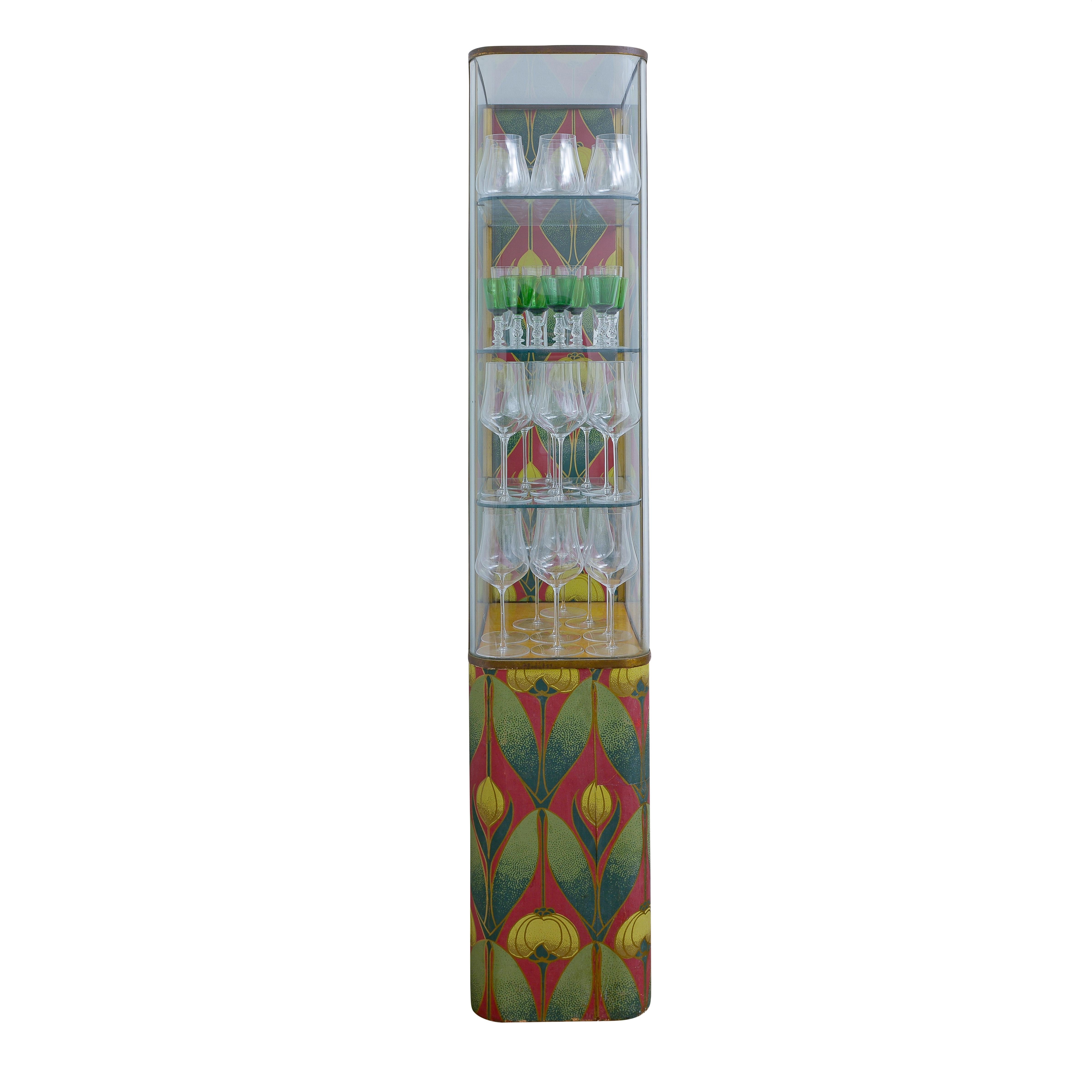 Llamativa vitrina Art Decó con marco de latón, paneles de cristal curvados y laterales con atrevidos dibujos en rojo, verde y dorado. La vitrina ofrece tres estantes de cristal para exponer dentro de una forma vertical alta, mostrando las líneas