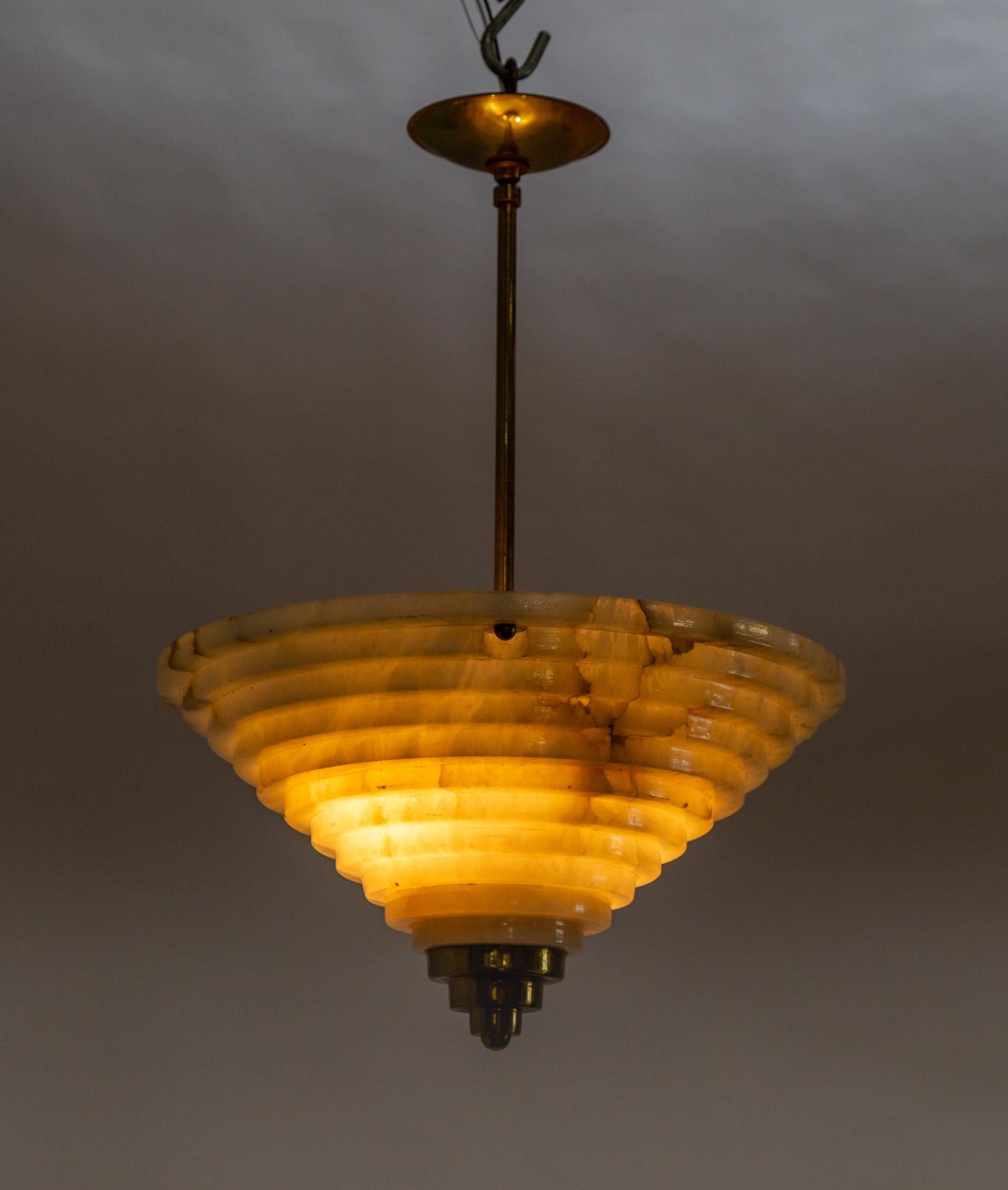 An Art Deco pendant light with a carved alabaster shade in an inverted stepped cone shape from a central patinated brass stem. The alabaster has a complex pattern of natural dark veining and is accented with a stepped brass point. It looks beautiful