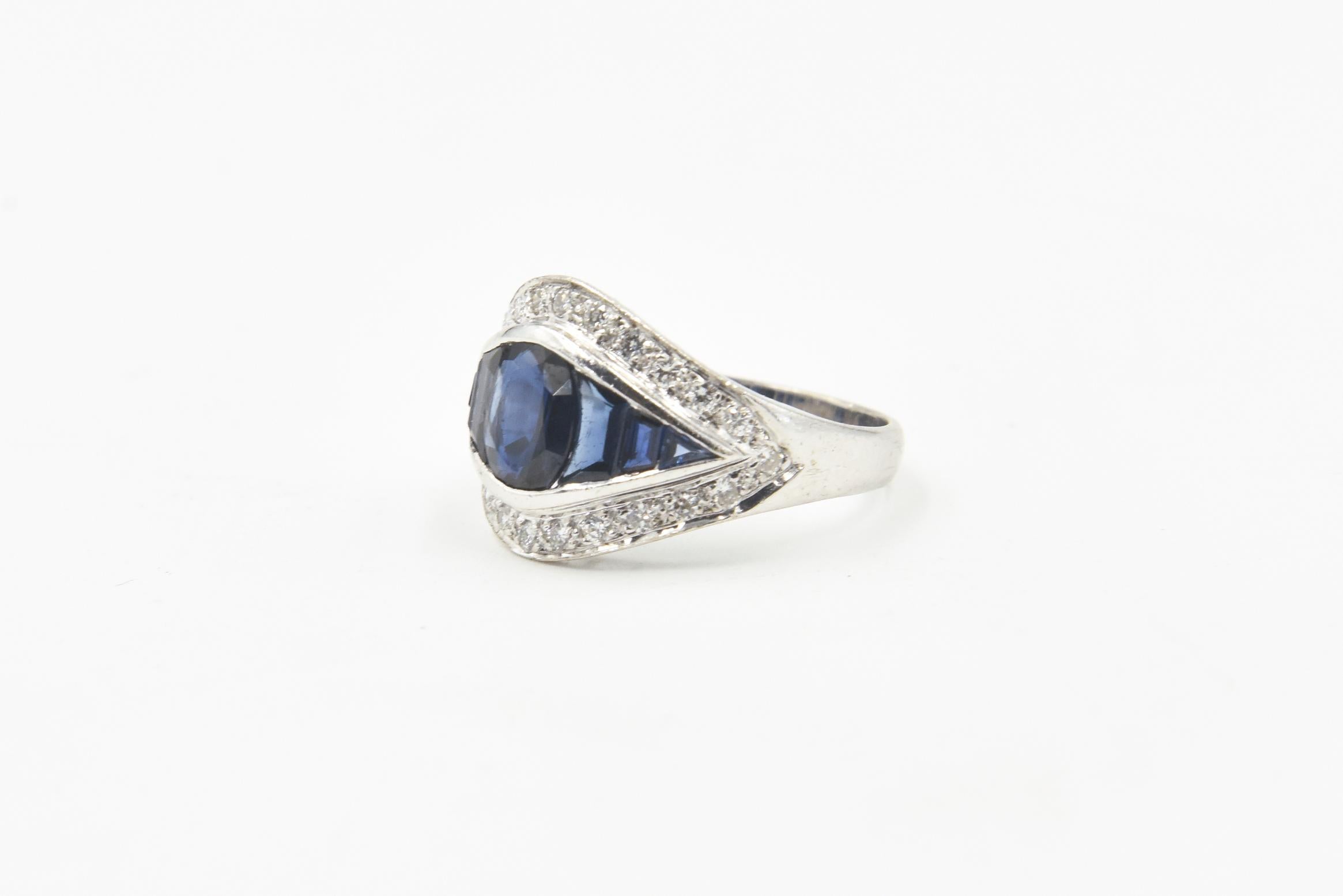 Art Deco-style cocktail ring featuring an oval faceted sapphire set beside graduating channel-set sapphires within a diamond frame. The mounting is 18k white gold.  
Marked: 18K.
 Size: US 6 3/4; can be sized.