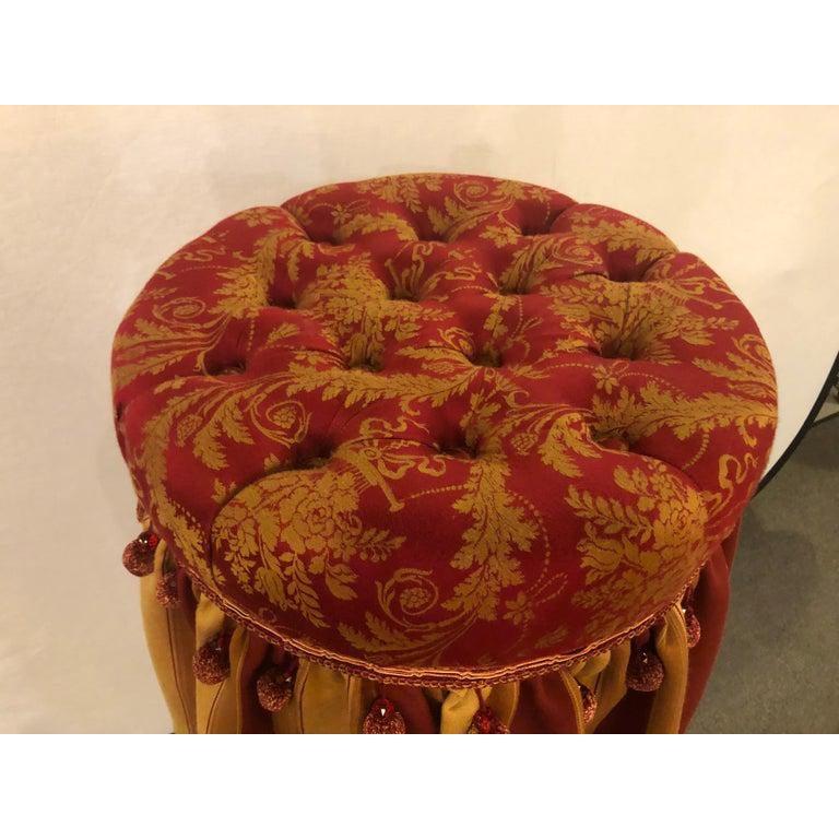 A Hollywood Regency upholstered tufted red and gilt decorated ottoman or footstool. This spectacular footstool or ottoman pouf is simply breathtaking. The finely upholstered piece with a tufted top and decorative base with stripped design and tassel