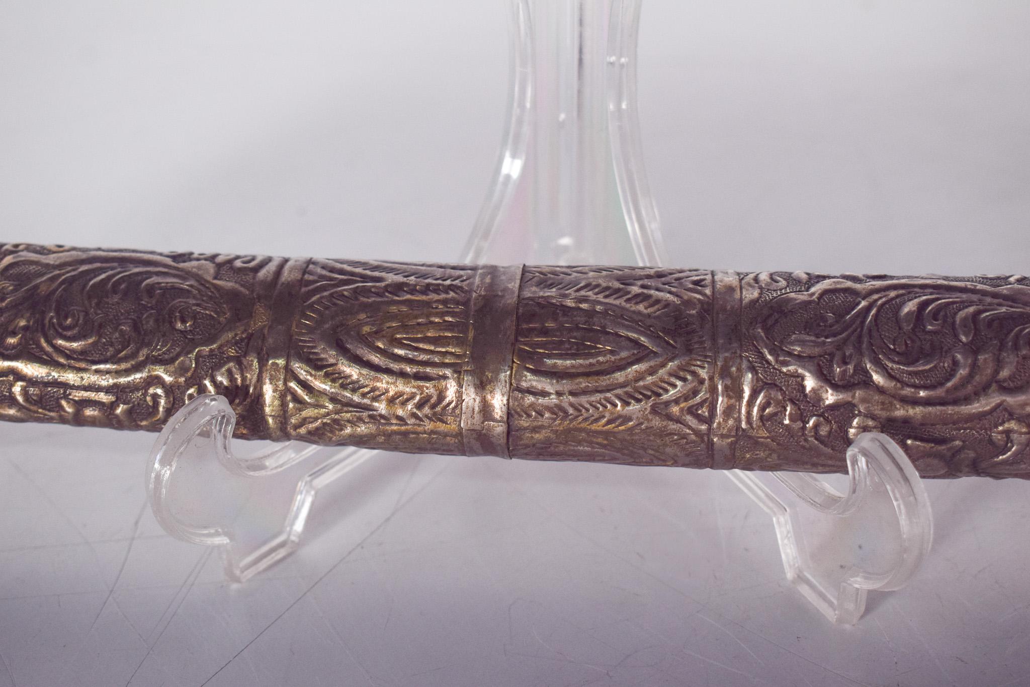 Poignard en argent décoré et repoussé. La lame porte des inscriptions et de petits motifs gravés, tandis que la poignée et le fourreau sont décorés de reliefs détaillés, peut-être inspirés de motifs traditionnels. Le fourreau s'adapte parfaitement à