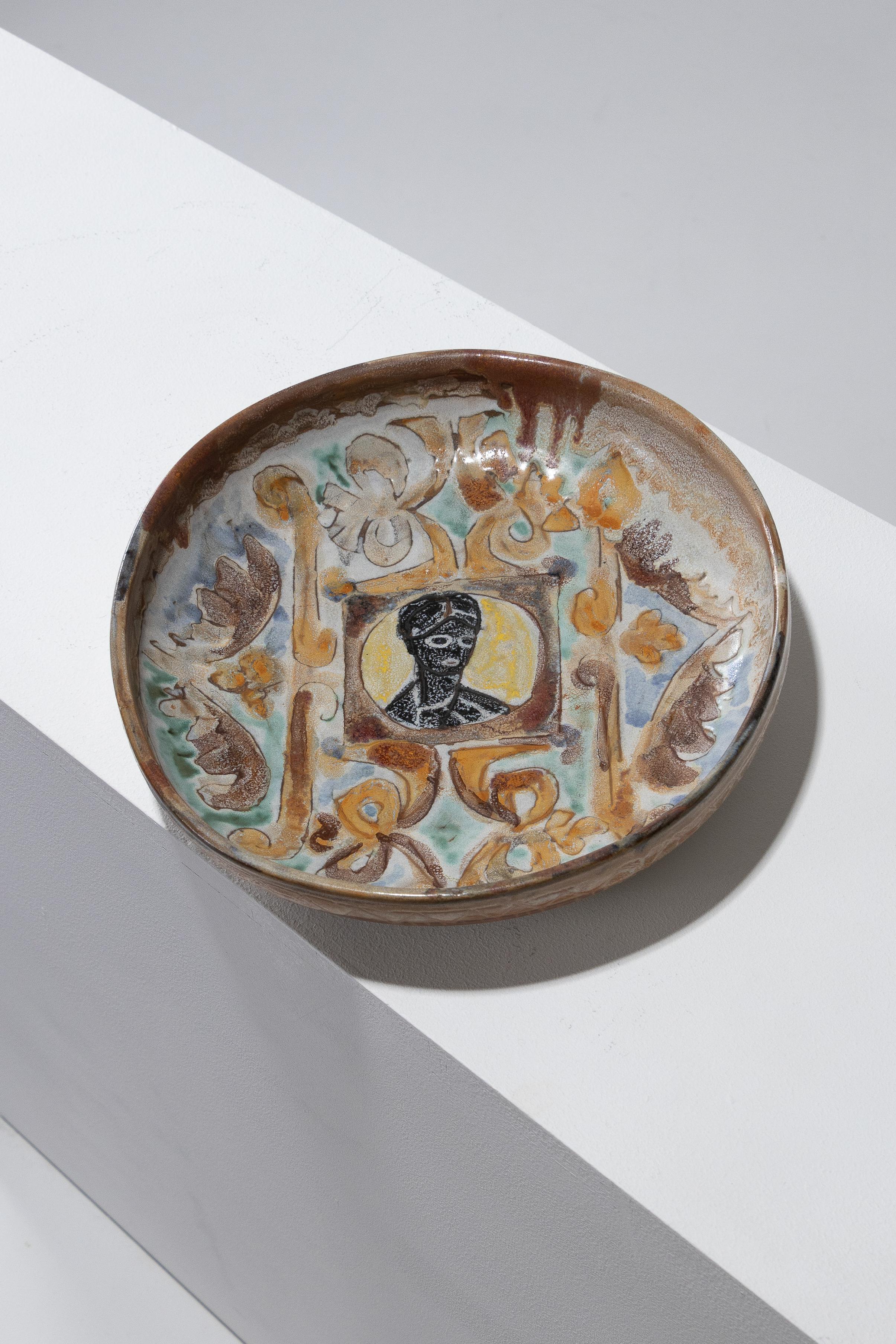 Placa de cerámica de aro alto fabricada en Vallauris en la década de 1950. El centro presenta una viñeta de una figura pintada en negro, rodeada de volutas policromadas sobre un fondo azulado. Firmado por el artista bajo la base. En excelente