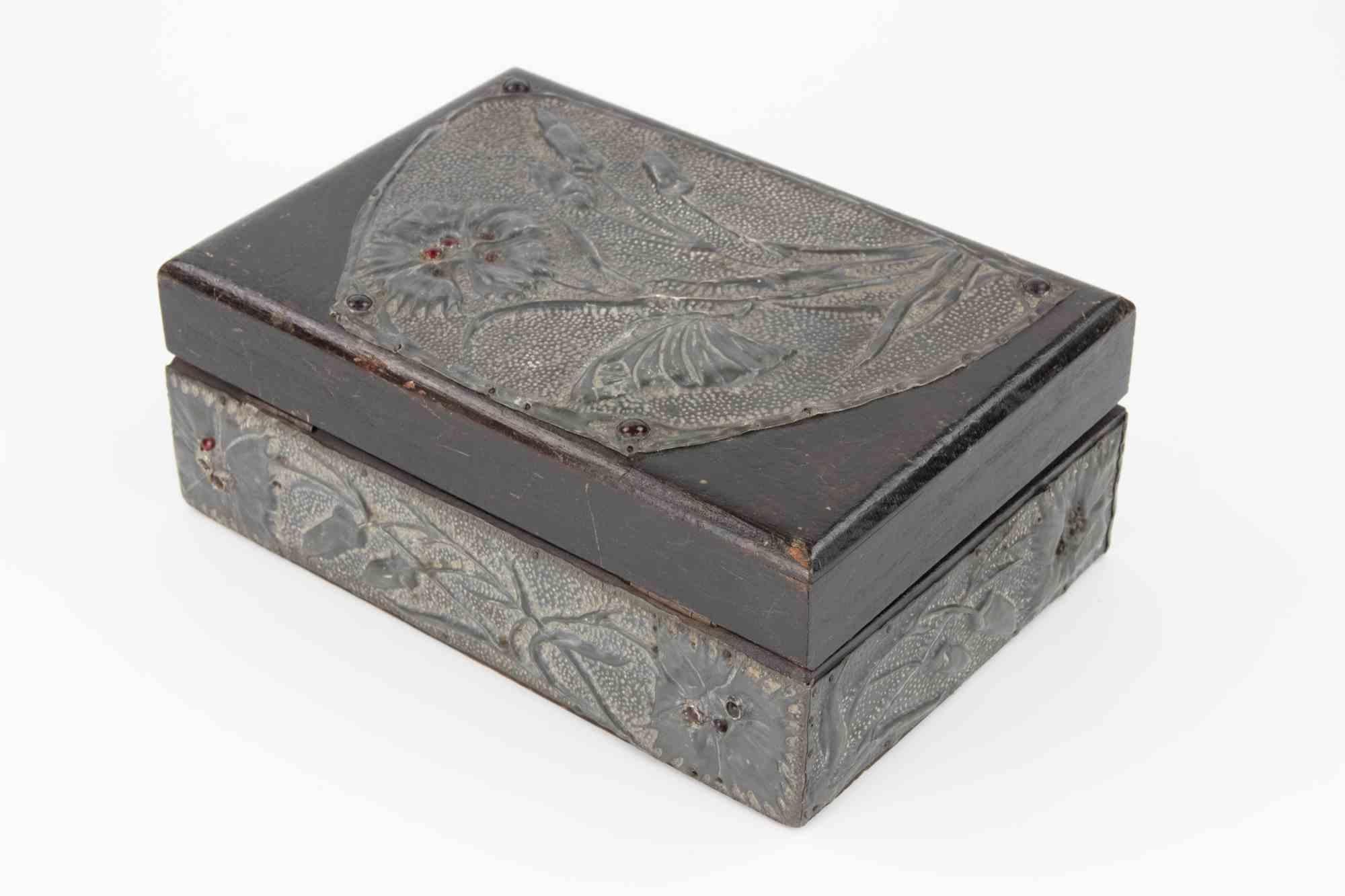 Uno scrigno rettangolare in legno con coperchio a cerniera, ornato da pannelli in peltro repoussé lavorati a basso rilievo. La decorazione raffigura fiori stilizzati, foglie e una farfalla tra il fogliame, il tutto su un fondo strutturato. I bordi