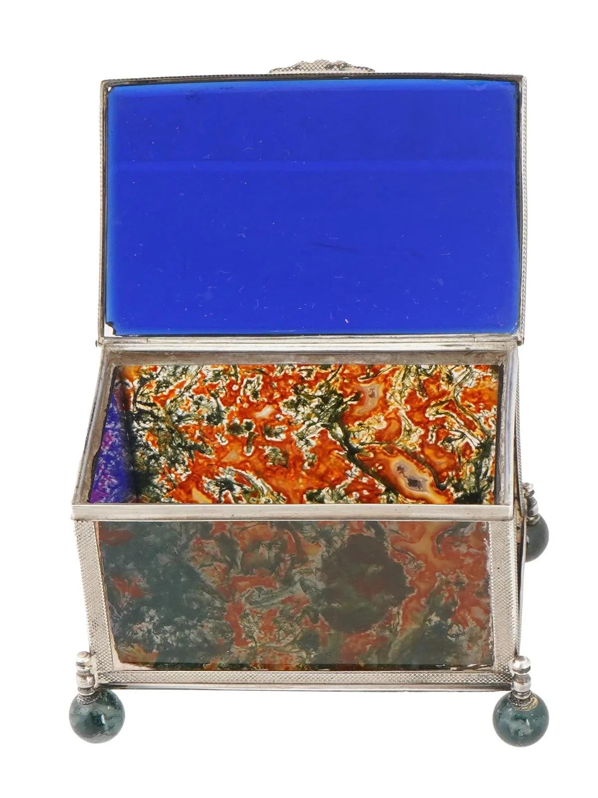 A decorative glass and moss agate box with a 925 sterling silver frame. Two sides of the box feature panels of moss agate, showcasing a mix of brown, red, and green hues. The lid and other sides is made of translucent glass. The box rests on round