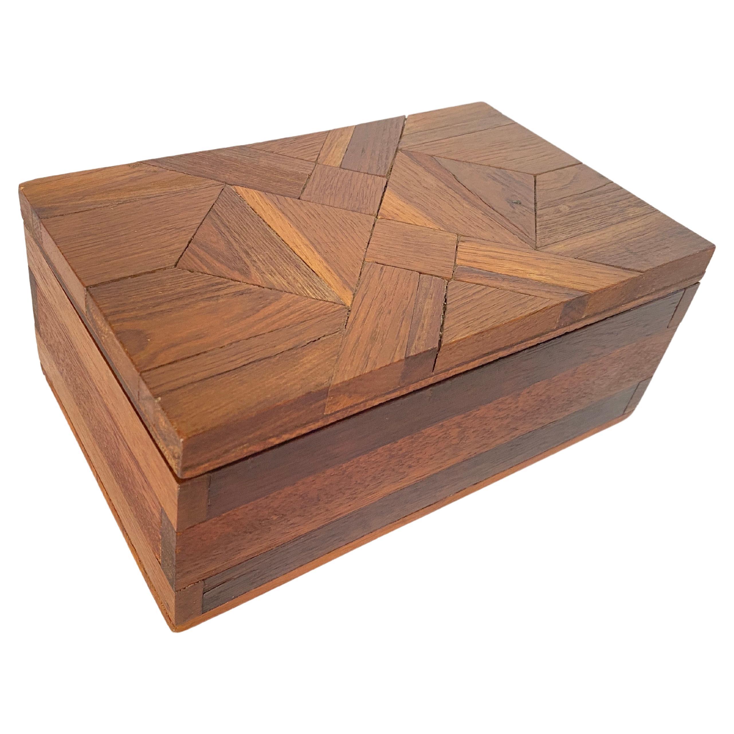 Dieses Kästchen ist ein Schmuckkästchen oder ein dekoratives Kästchen. Es wurde in den 1970er Jahren in Frankreich hergestellt. Der Deckel besteht aus einem schachbrettartigen Rechteck aus Holz. Die Farbe ist braun.
