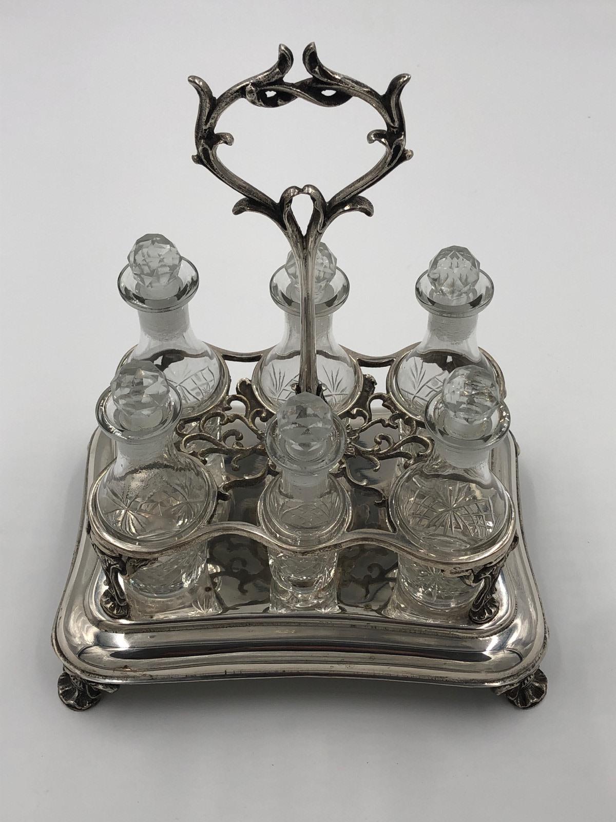 Antico set di 6 bottiglie, placcato in argento, con 5 bottiglie della stessa dimensione e una più piccola al centro, tutte con tappi originali. Maniglia di trasporto ornata e piedi a foglia cabriole. Buone condizioni.
