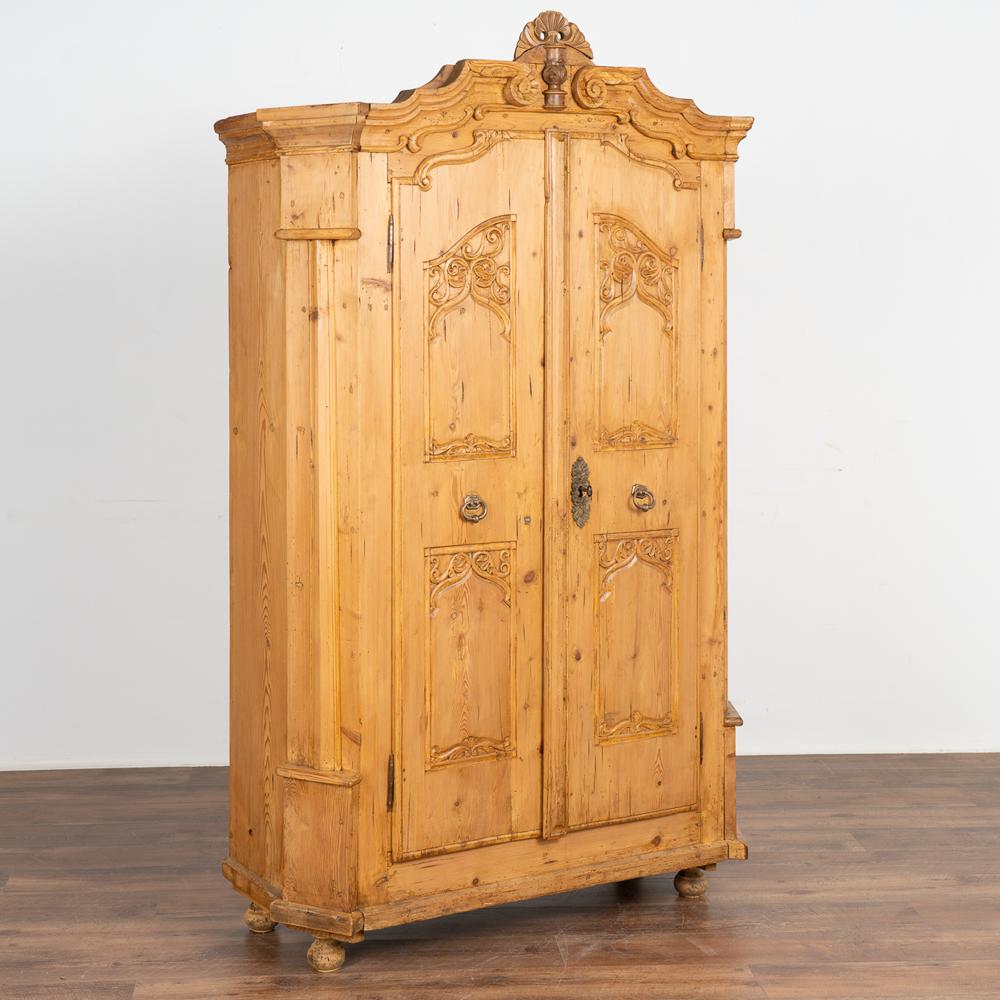 Decorative carving in the panels and crown add a sophisticated touch to this small two door pine armoire with an aged patina from years of use.
Refer to interior photos to appreciate how it is divided to allow for tall items on the left (currently