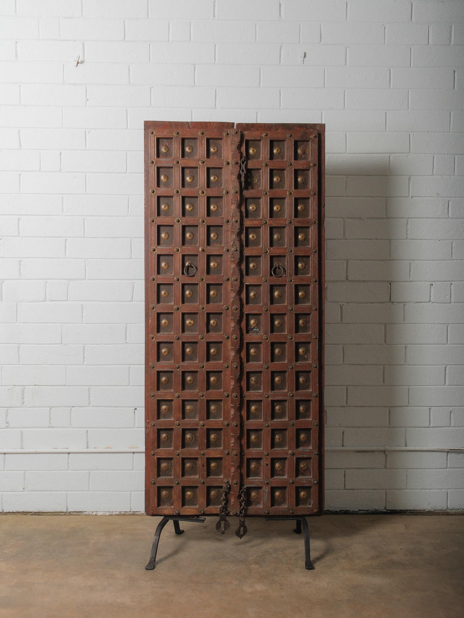A striking architectural fragment presented as sculptural décor, this carved wooden door is mounted on a custom iron stand, transforming a once-functional element into an expressive design object. Its richly weathered surface reveals traces of