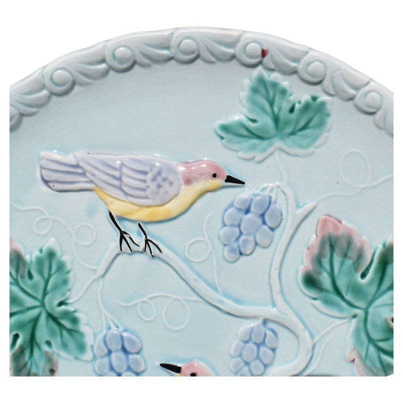 A pretty pale blue majolica bird motif plate. This decorative plate would be fabulous hung on a wall or displayed in a china cabinet. Raised textured light blue birds perch on blue branches and are surrounded by green and pink leaves. The outer edge