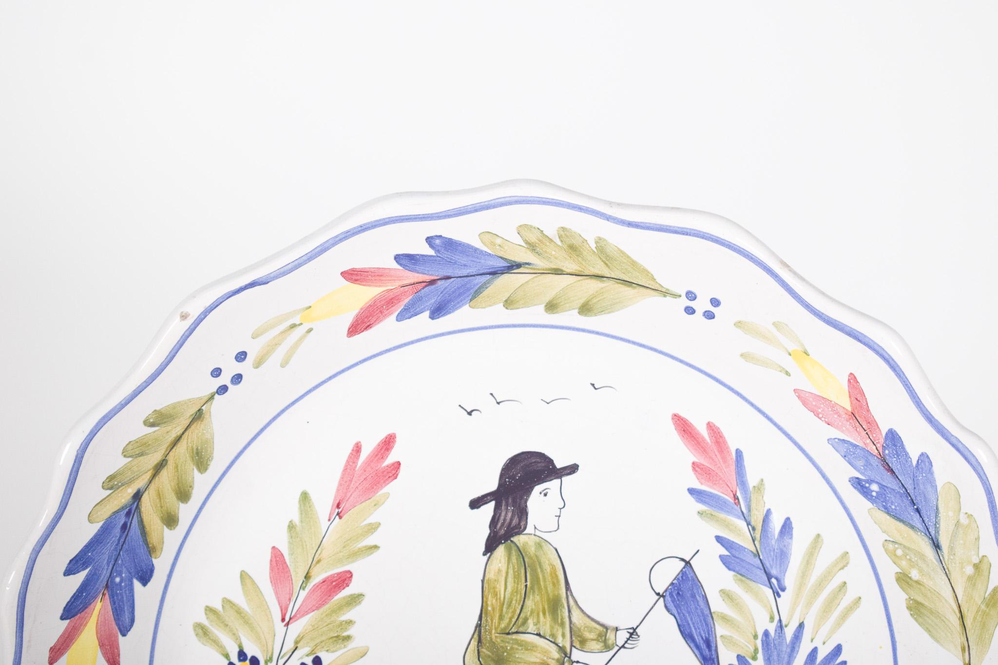 Plat en céramique avec pêcheur. Au centre se trouve la figure d'un homme portant un chapeau noir, une chemise verte et un short bleu, tenant un filet de pêche avec un manche. Autour de la figure se trouvent des branches peintes de feuilles vertes,