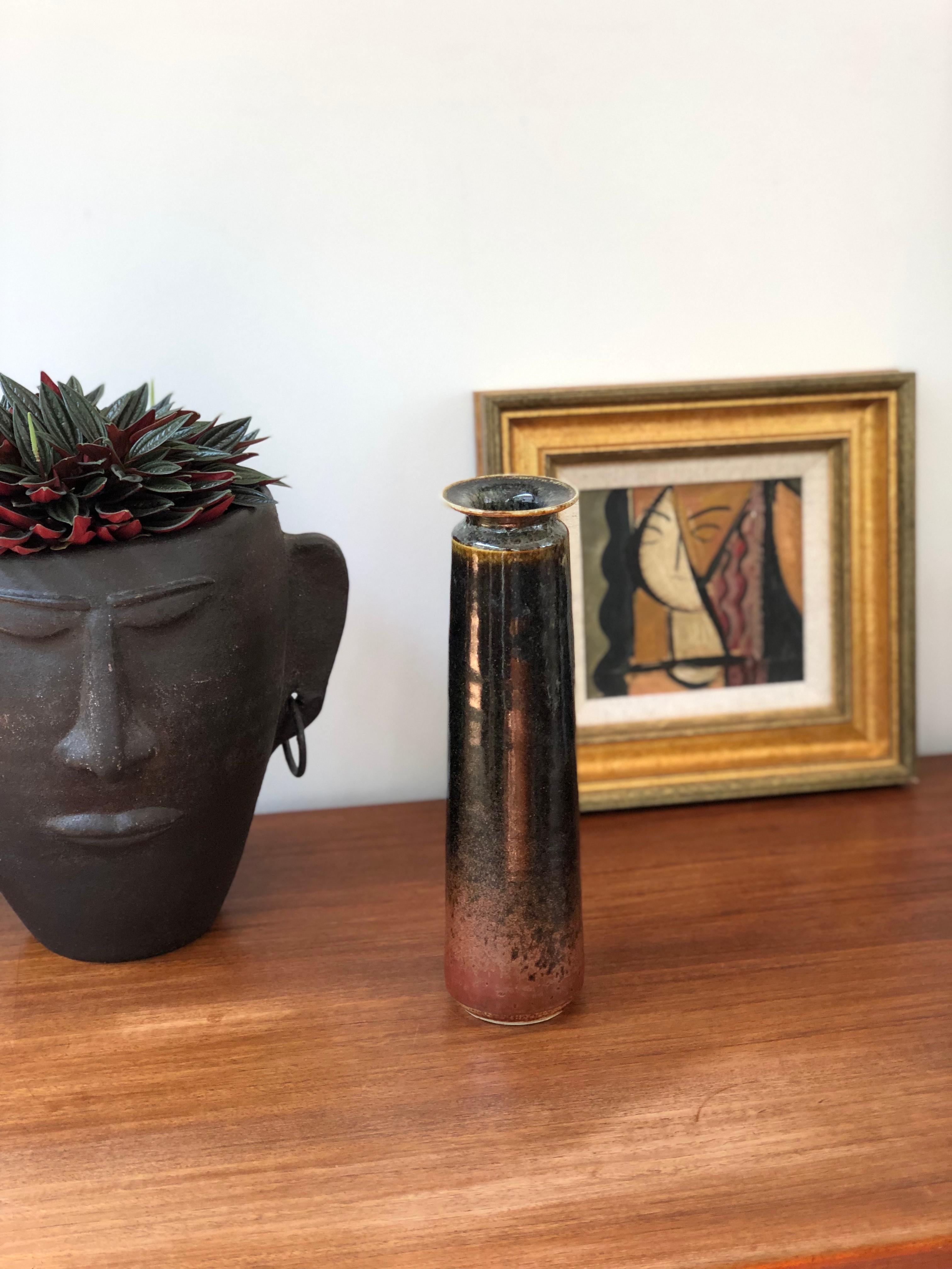 Decorative elongated ceramic flower vase (circa 1960s). This mid-century flower vase is cylindrical in shape and stylishly glazed with a sheen in a dark brown interrupted with lighter highlights and much lighter coloring near the base. At the top,