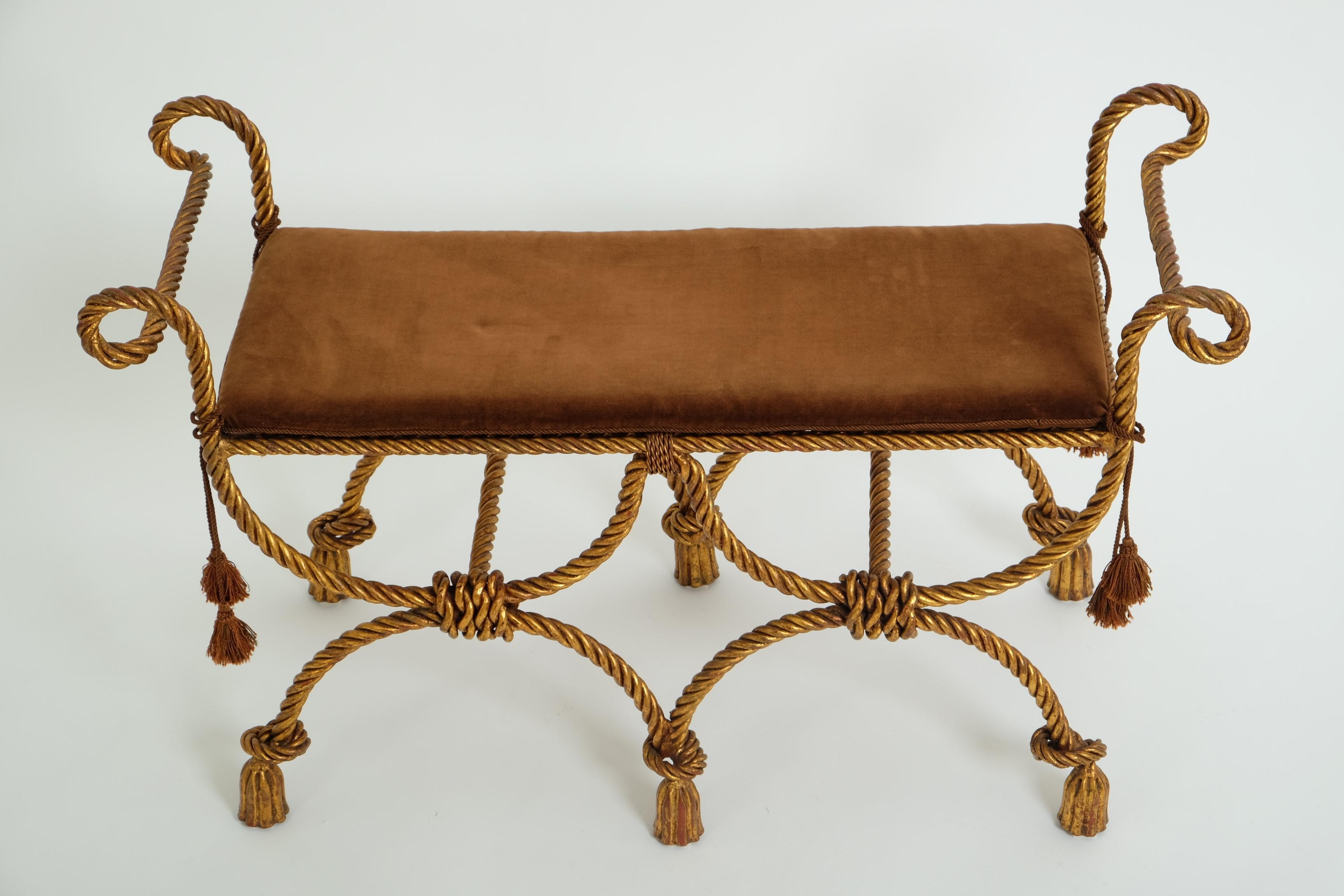 Banc décoratif Hollywood Regency en fer forgé doré, Italie années 1950.

Lourde banquette en fer doré avec une double armature en forme de curule faite de cordes stylisées et de pieds à glands. L'assise perforée est complétée sur les côtés par des