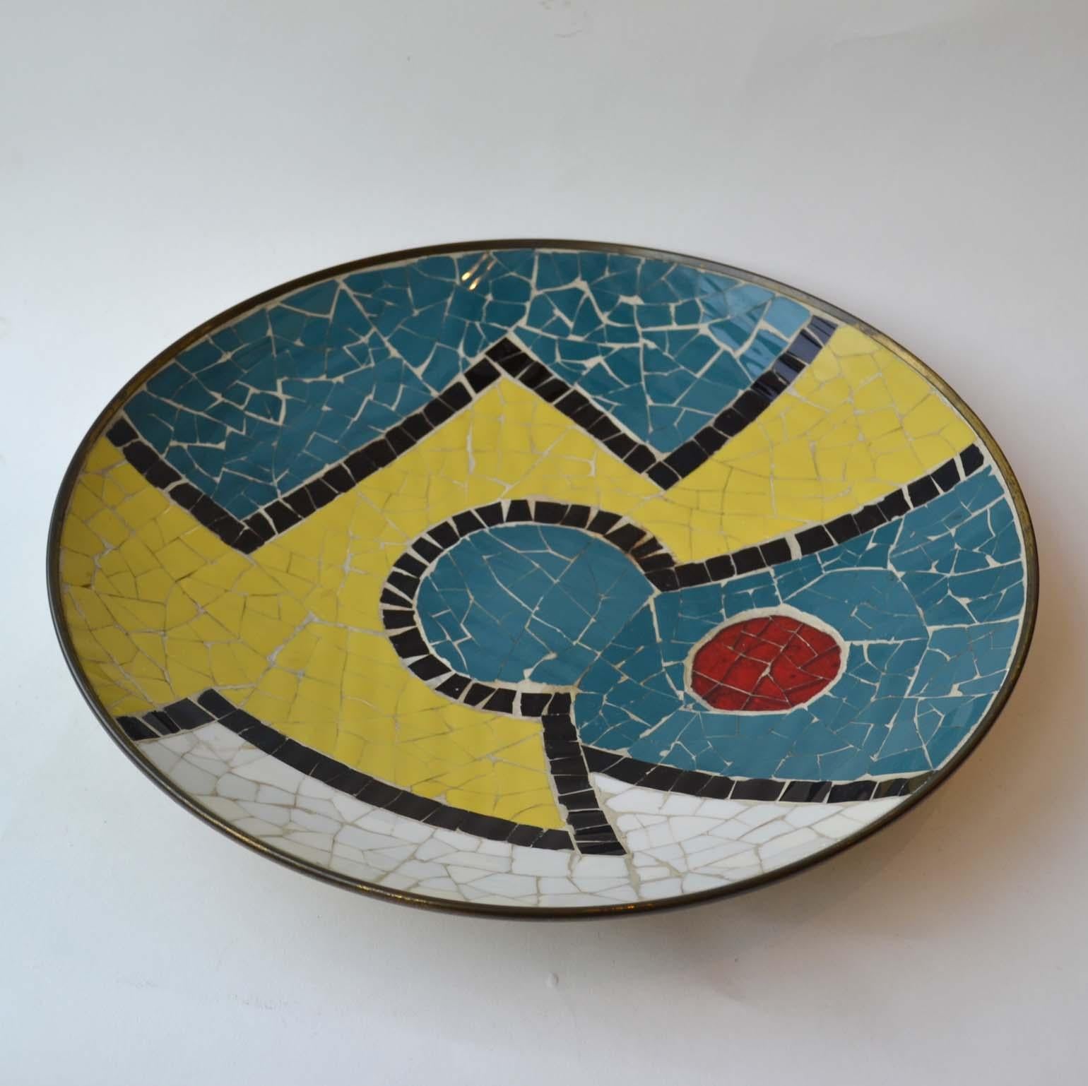 Multi-color mosaic charger made in the 1960s designed to hang on the wall. The glass mosaic is laid in blocks of colour with strong black borders on to a brass plate with a raised foot.