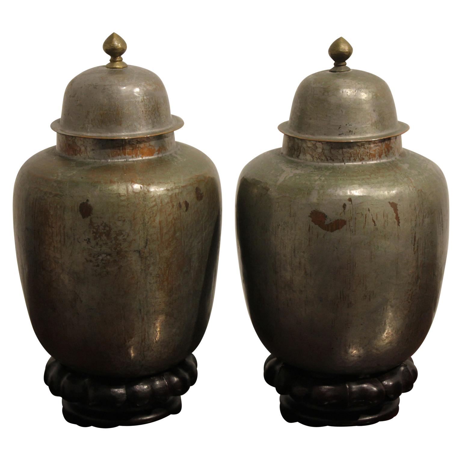 Gilt copper over tin urn-shaped vessels that sit on a decoratively carved wooden pedestal. The vessels are sold as a pair and have detachable lids.
Dimensions are of one urn with the pedestal.
 