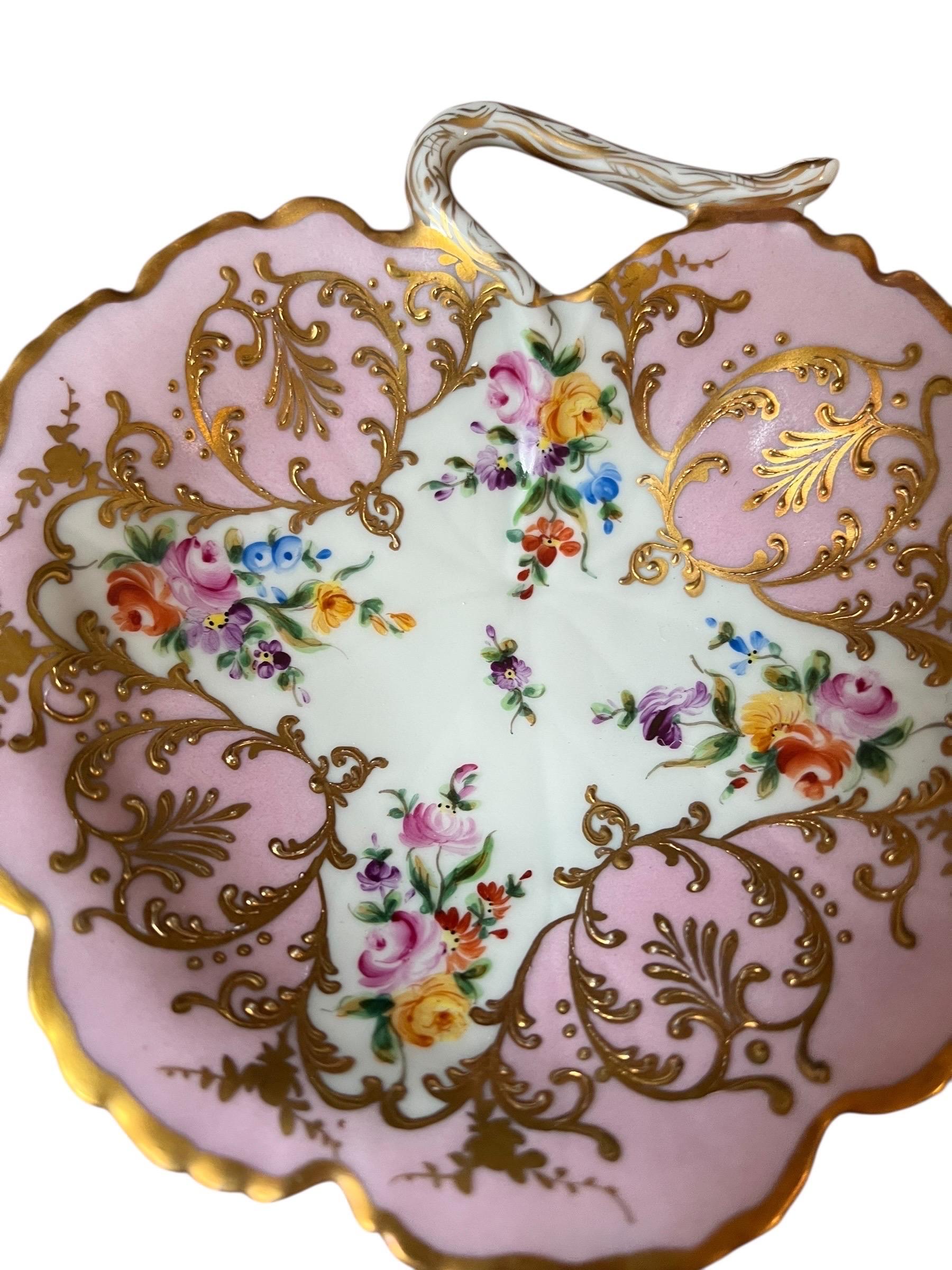 Un élégant plat en porcelaine de Paris, finement travaillé dans une forme fluide d'inspiration rococo. La pièce est rehaussée d'un riche décor polychrome de guirlandes florales et d'arabesques dorées appliquées à l'or fin, dans un style typiquement