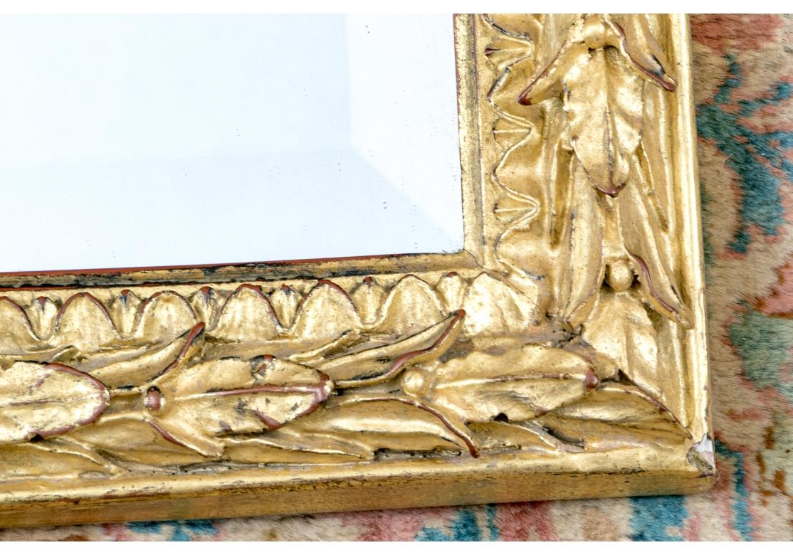 Miroir rectangulaire biseauté de style rococo avec un cadre doré orné d'un fronton à couronnes de laurier feuillues menant à des vignes florales sur un périmètre extérieur de feuillage texturé et une garniture intérieure de fins pétales en gradins.
