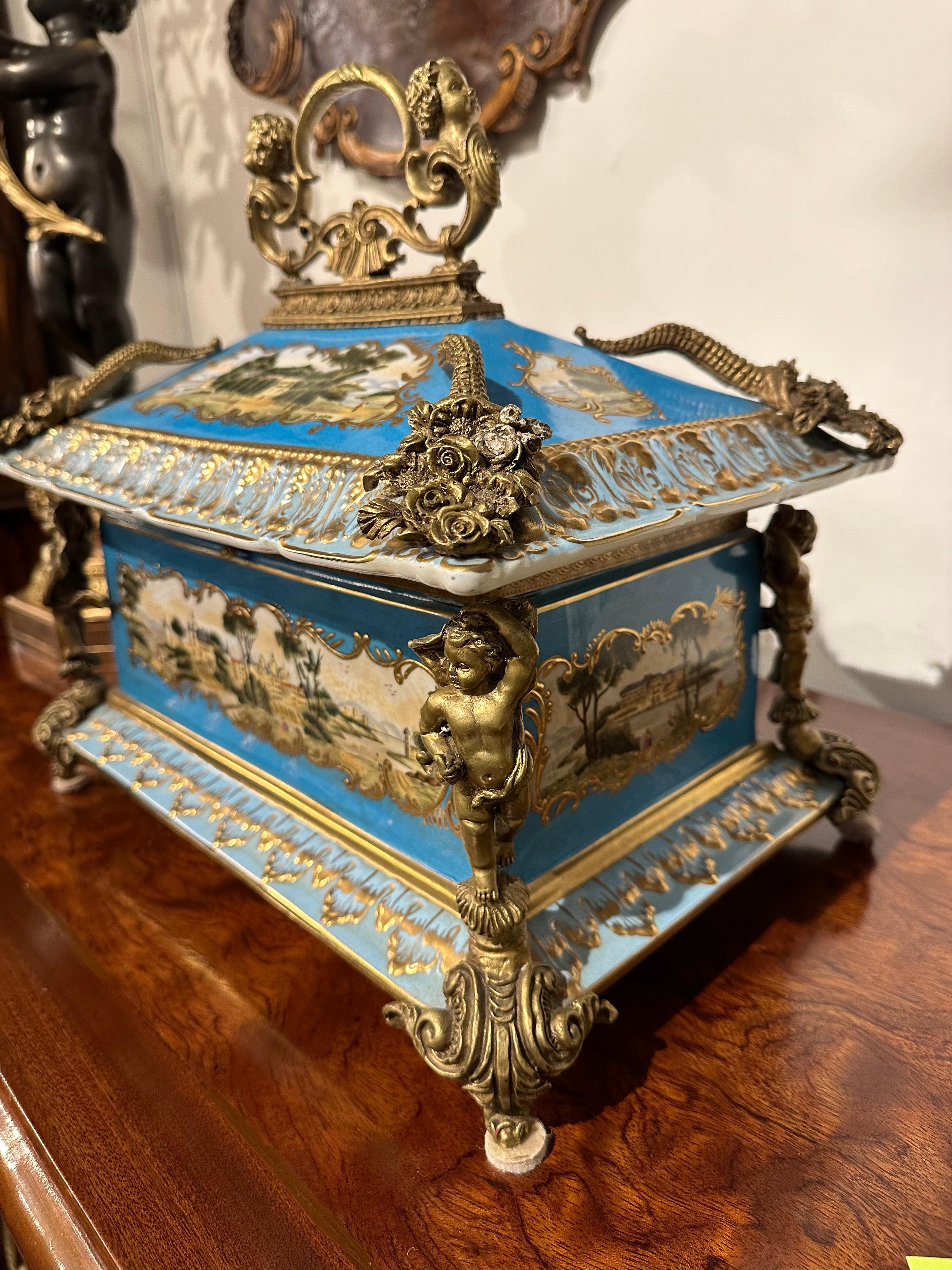 Elegant and highly decorative porcelain box with exquisite painted scenes and bright colours on a sky blue background depicting a landscape and country house. Internally there is a delicate painting of a floral bouquet in vivid colours. The side
