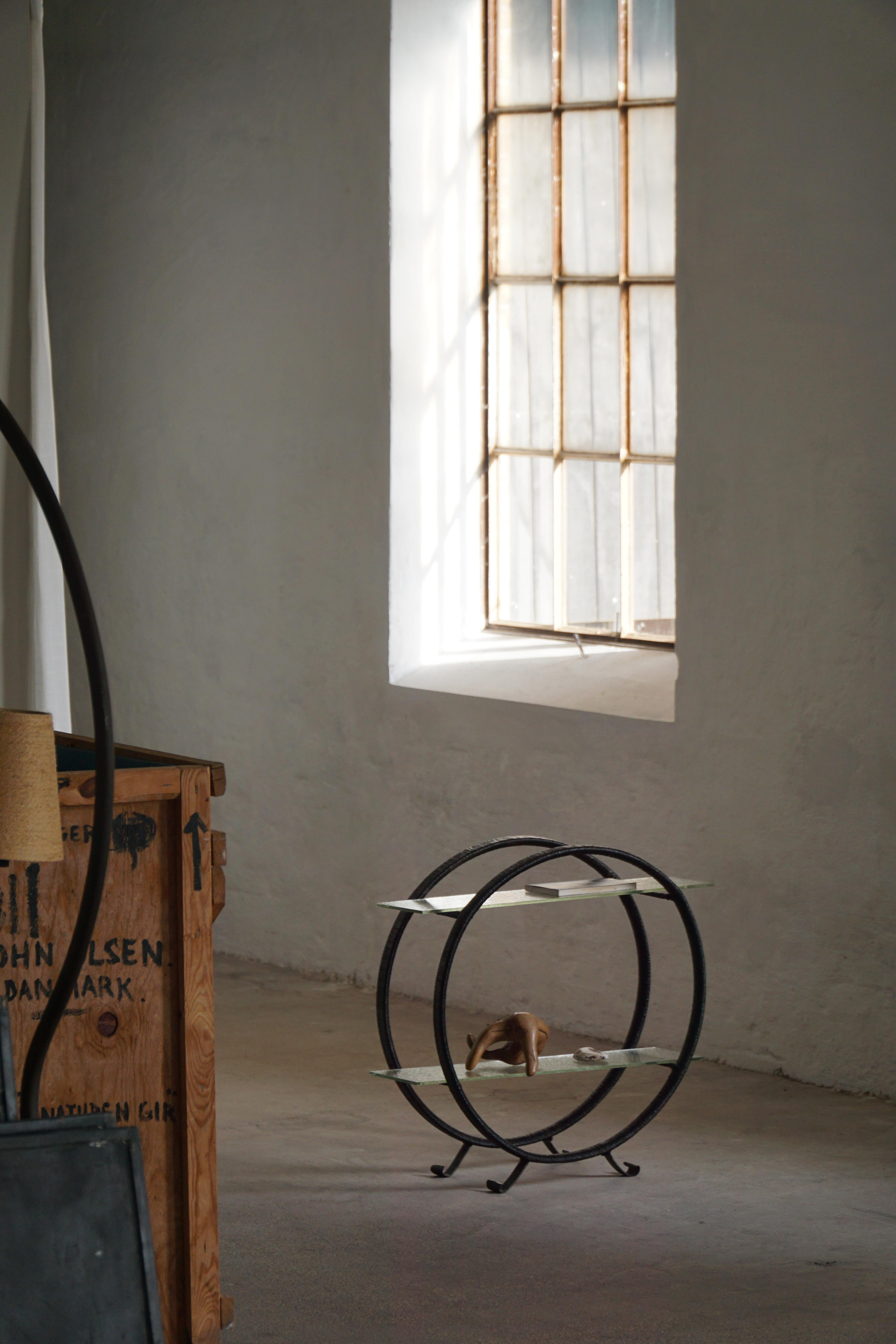 Un piccolo scaffale decorativo scandinavo elegante e minimalista, caratterizzato da due telai circolari in metallo sostenuti da quattro gambe metalliche elegantemente ricurve. L'unità comprende due ripiani in vetro che conferiscono un aspetto